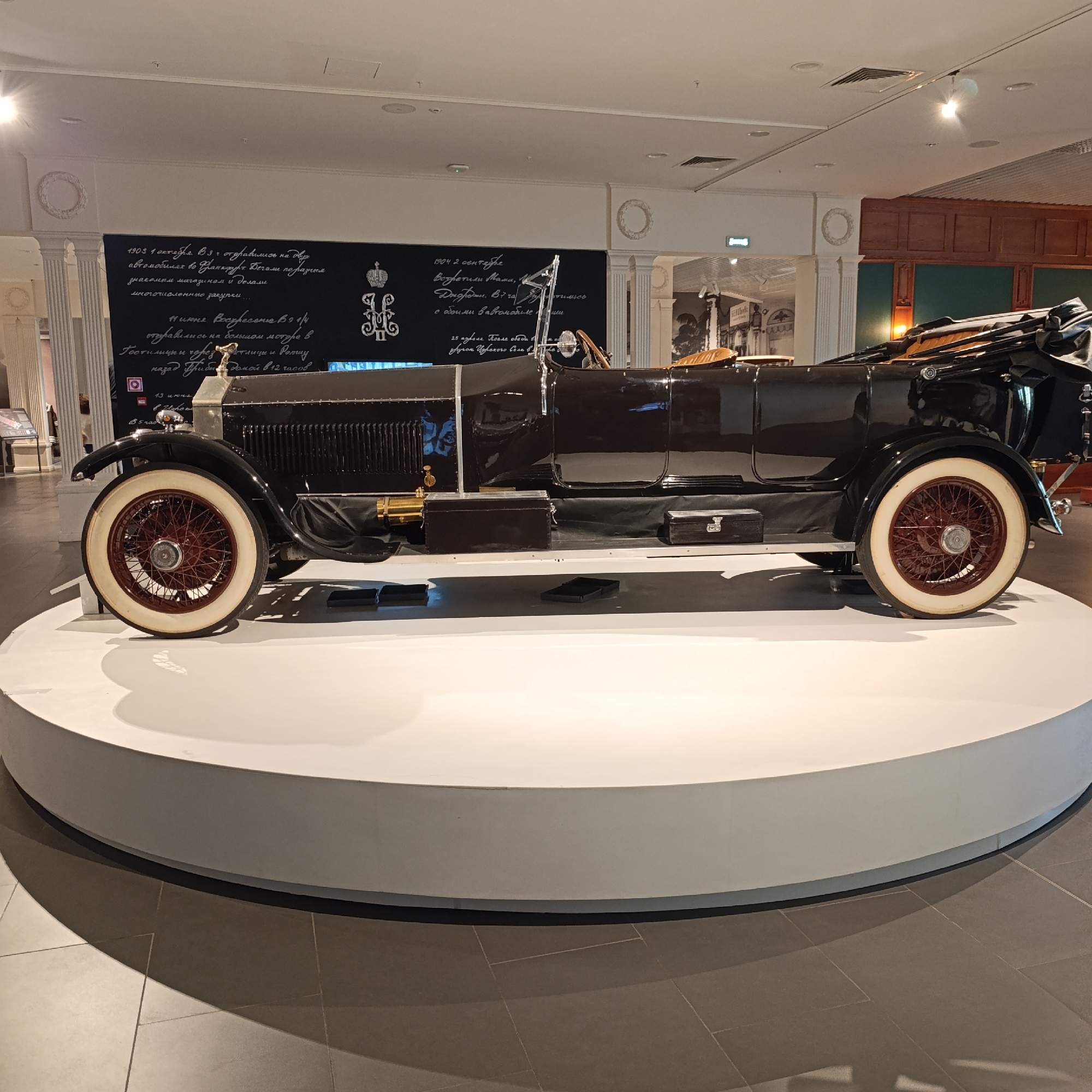 The image shows a vintage black car on a white circular platform inside a building. The car has large white-walled tires with red rims. Behind the car is a wall with text written on it in a white script. The setting appears to be a museum. The car is a luxury model, possibly a Rolls Royce or similar high-end vintage vehicle, featuring a large radiator grill and other period-specific design elements. Based on the visual elements, the location is likely a museum in Verkhnyaya Pyshma, Russia, known for its large collection of vintage vehicles.