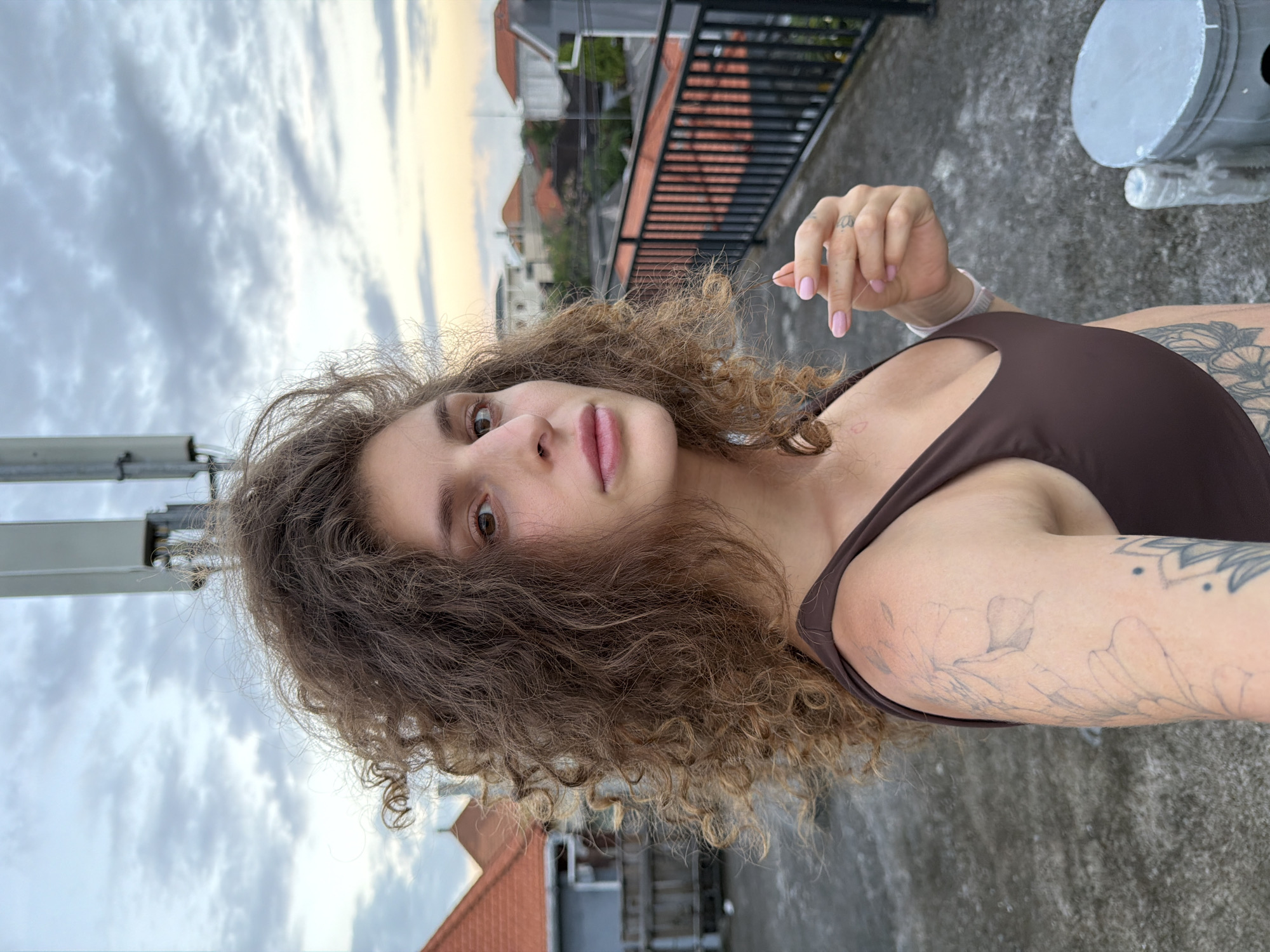 A young woman with light skin and curly brown hair poses outside on a concrete rooftop. She wears a brown top. Her left arm shows tattoos. The background shows a cloudy sky, rooftops of buildings, and a metal railing. The location is identified as North Kuta, Indonesia.