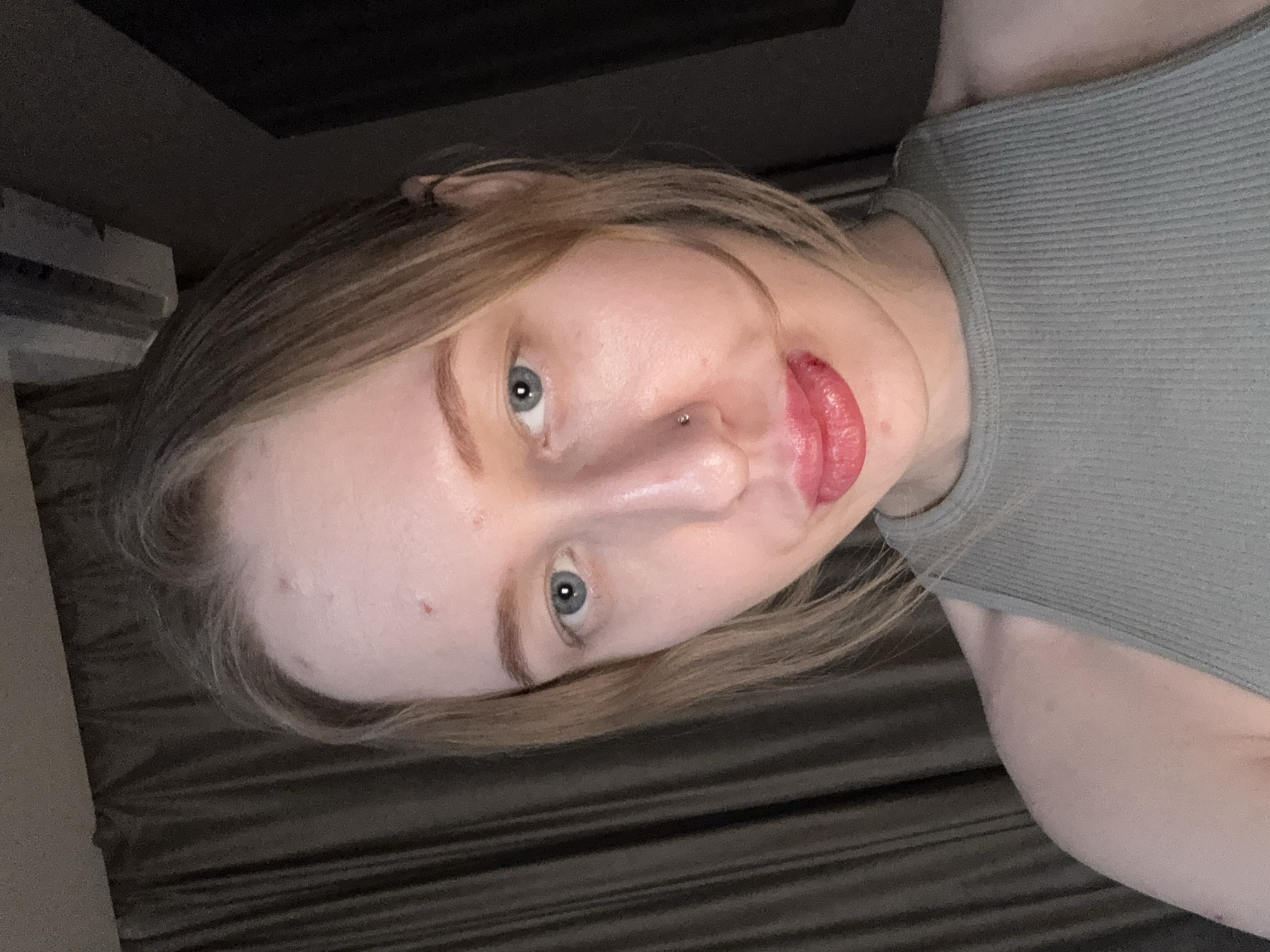 A light-skinned person with blue eyes and blonde hair poses for a selfie. They are wearing a ribbed, grayish-green sleeveless top. A nose piercing is visible. The background is indistinct, showing a dark surface and what appears to be a partially visible window frame. No information suggests the location is Rostov-on-Don, Russia.