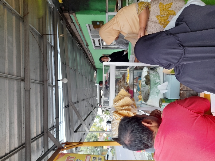 A food stall is set up under a metal awning. Customers are gathered around, including a person in a hijab, a woman in a light brown patterned top, and a person in a red shirt. A glass display case holds various food items, including what appears to be crackers or chips. A tissue box sits on the counter. A person in a black jacket stands in the background near a green building.