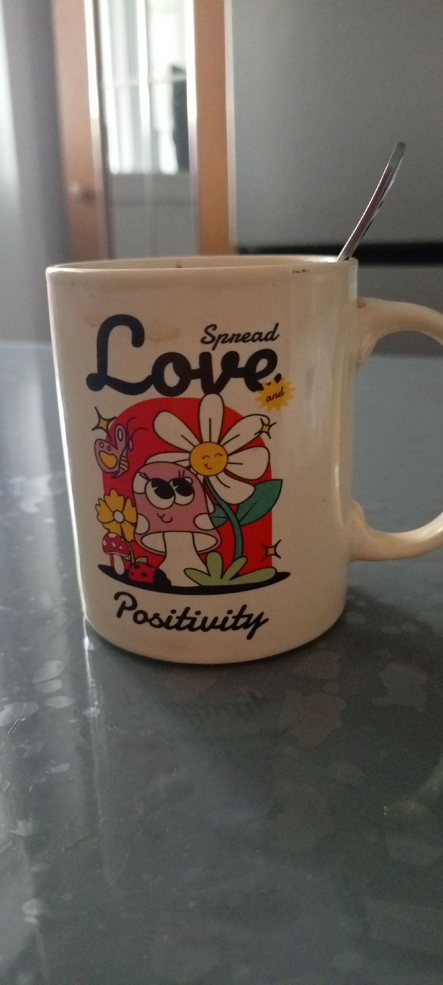 A close-up, eye-level, vertical shot shows a cream-colored ceramic mug with a spoon inserted. The mug has a graphic design featuring the words "Spread Love and Positivity" written in black script. The design also includes cartoonish illustrations of mushrooms, flowers, and a butterfly. The mug is placed on a dark, reflective surface, possibly a table. In the blurred background, a doorway with glass panes and wooden trim is visible. This scene is located in Tarasovka, Russia.