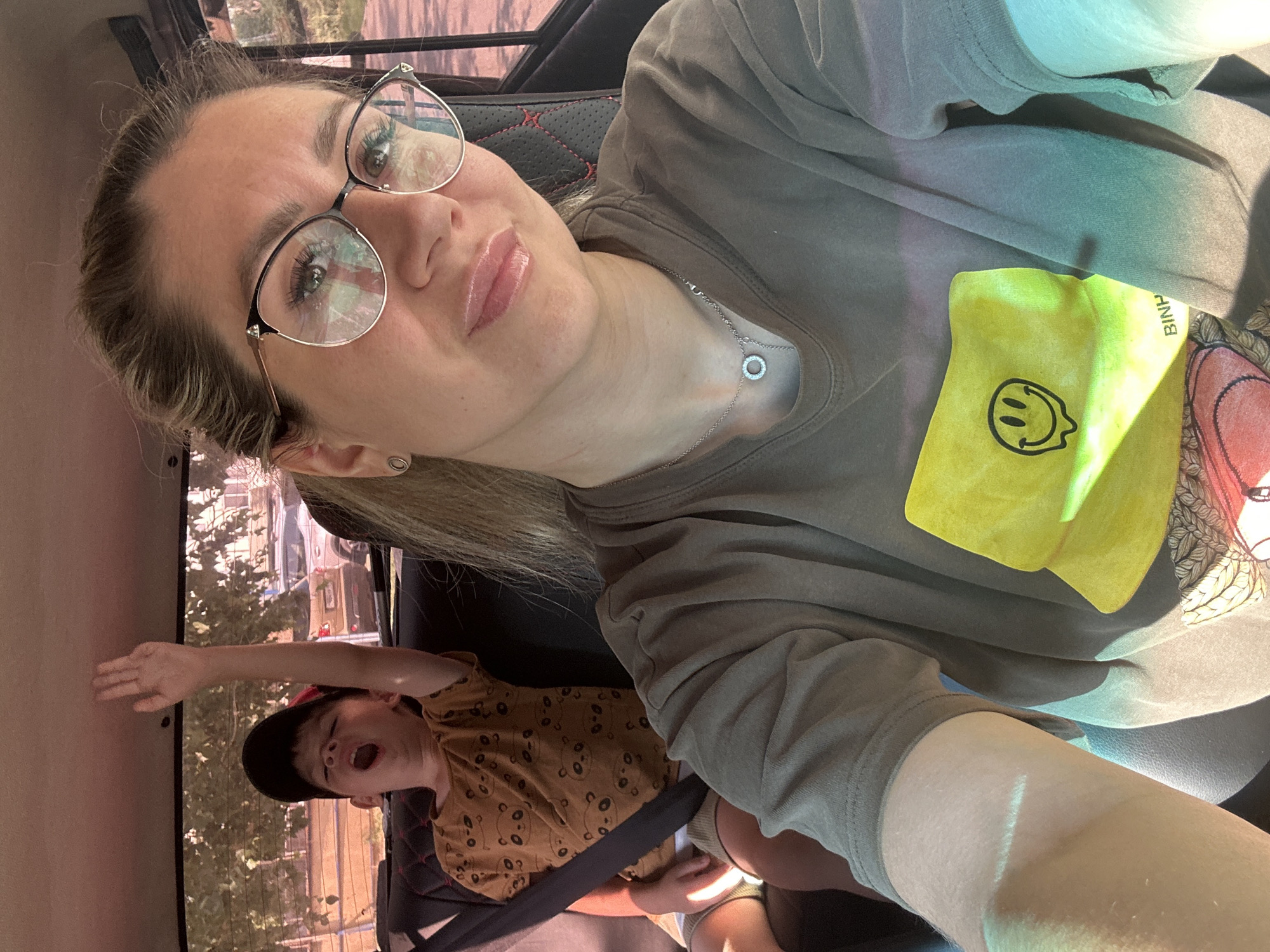 In the image, a woman with blonde hair and glasses is taking a selfie from the front passenger seat of a car. She is wearing a gray t-shirt with a yellow smiley face graphic. In the back seat, a young boy wearing a baseball cap and a brown t-shirt with a panda pattern is visible. He has his mouth open and his right arm raised. The car appears to be parked or driving on a street in Novoaltaysk, Russia, with trees and other cars visible through the windows, suggesting an outdoor, daytime setting.