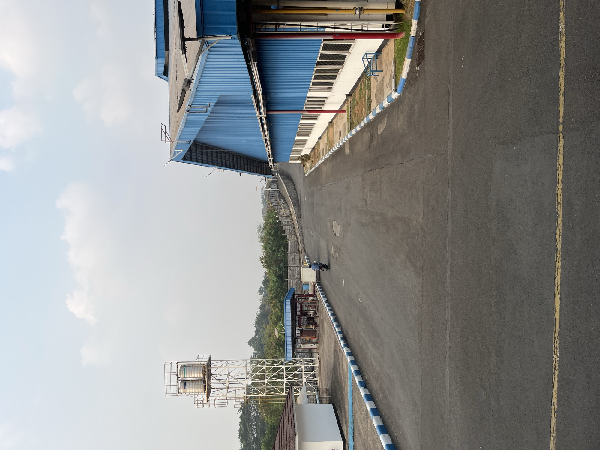 The image captures an industrial scene on a bright day with a hazy sky. A paved road leads into a facility with a large, blue, corrugated metal building on the right. This building features a row of windows along its lower half, and red and yellow pipes run vertically along its exterior. To the left of the road, there's a white building with a brown roof, a tall white water tower with two cylindrical tanks, and an open-sided structure housing industrial equipment and tanks. A single individual is riding a dark-colored motorcycle away from the viewer down the road. The road itself is marked with yellow lines and bordered by a curb with alternating blue and white stripes. In the background, a concrete wall and lush green trees are visible, suggesting a semi-rural or suburban industrial setting. The overall impression is one of a functioning industrial compound. The location is identified as the city of Pelang, Indonesia.