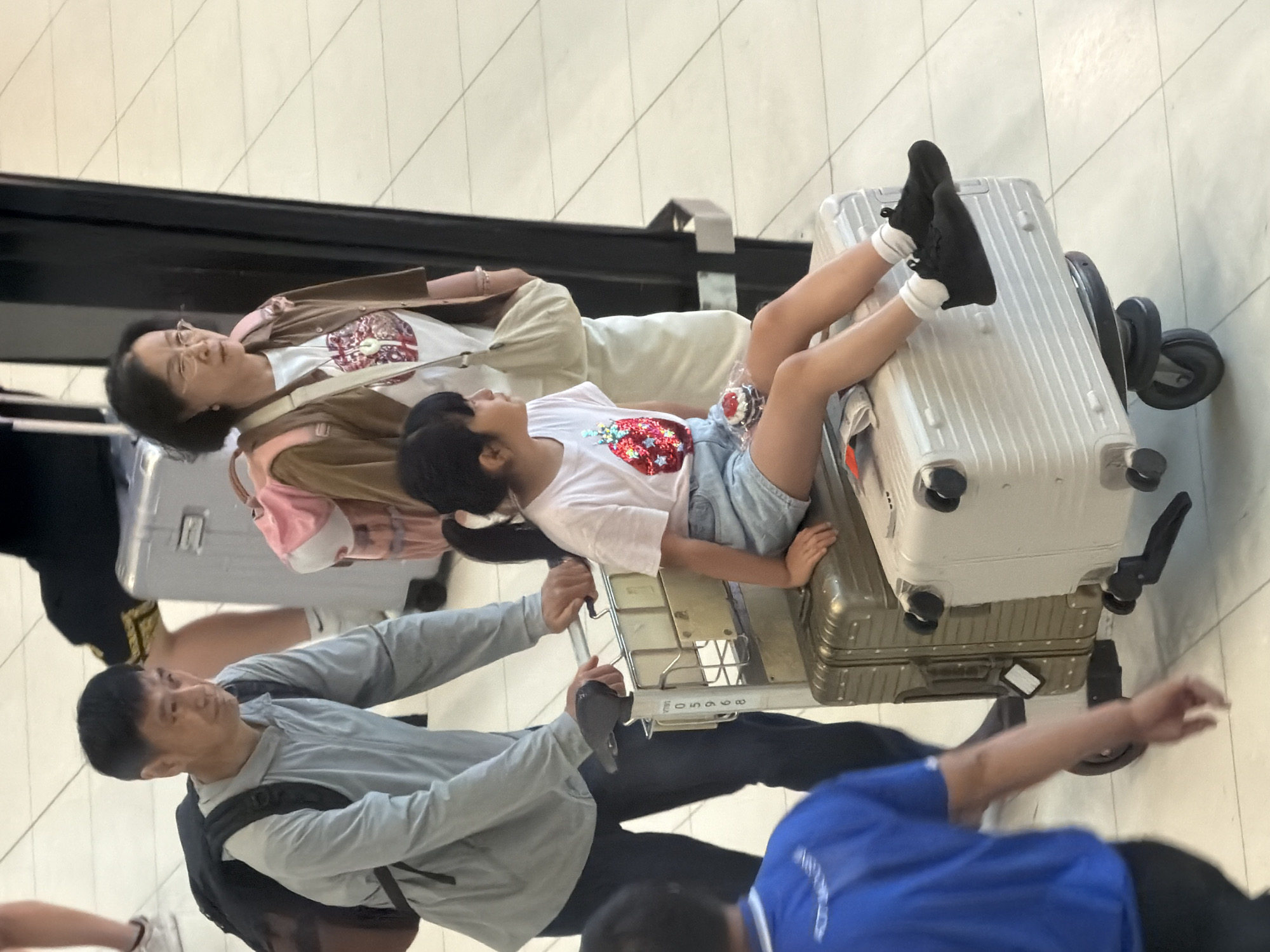 In the city of Nong Prue Subdistrict, Thailand, an overhead shot captures a family navigating an airport. A man in a grey hooded jacket and dark pants pushes a luggage cart laden with two suitcases. Seated on the top suitcase is a young girl wearing a white t-shirt with a sequined fruit design and denim shorts. She is holding a cupcake-like object. Behind her, a woman in a brown jacket and white t-shirt, wearing glasses and a pink backpack, stands near the luggage cart. Another person in a blue shirt is partially visible on the left, reaching towards the luggage. The environment is characterized by a tiled floor and the presence of airport infrastructure like luggage carts and tall dark structures.
