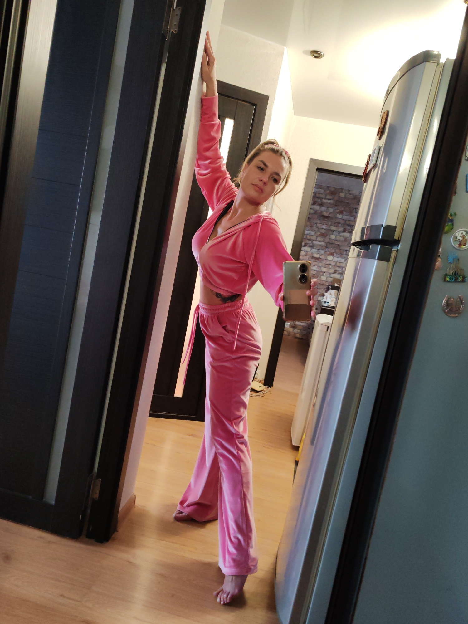 A woman is standing indoors, posing for a selfie in a pink velour tracksuit. She is positioned near a doorway with dark frames and paneling, holding a smartphone in her right hand. Her left arm is extended upwards, leaning against the doorway. A silver refrigerator is to her right, reflecting the scene and objects within the room. In the background, a hallway leads to a room with exposed brick walls. The location is likely in an apartment or house. It is not possible to determine if this is located in Petropavlovsk-Kamchatsky, Russia from the image alone.