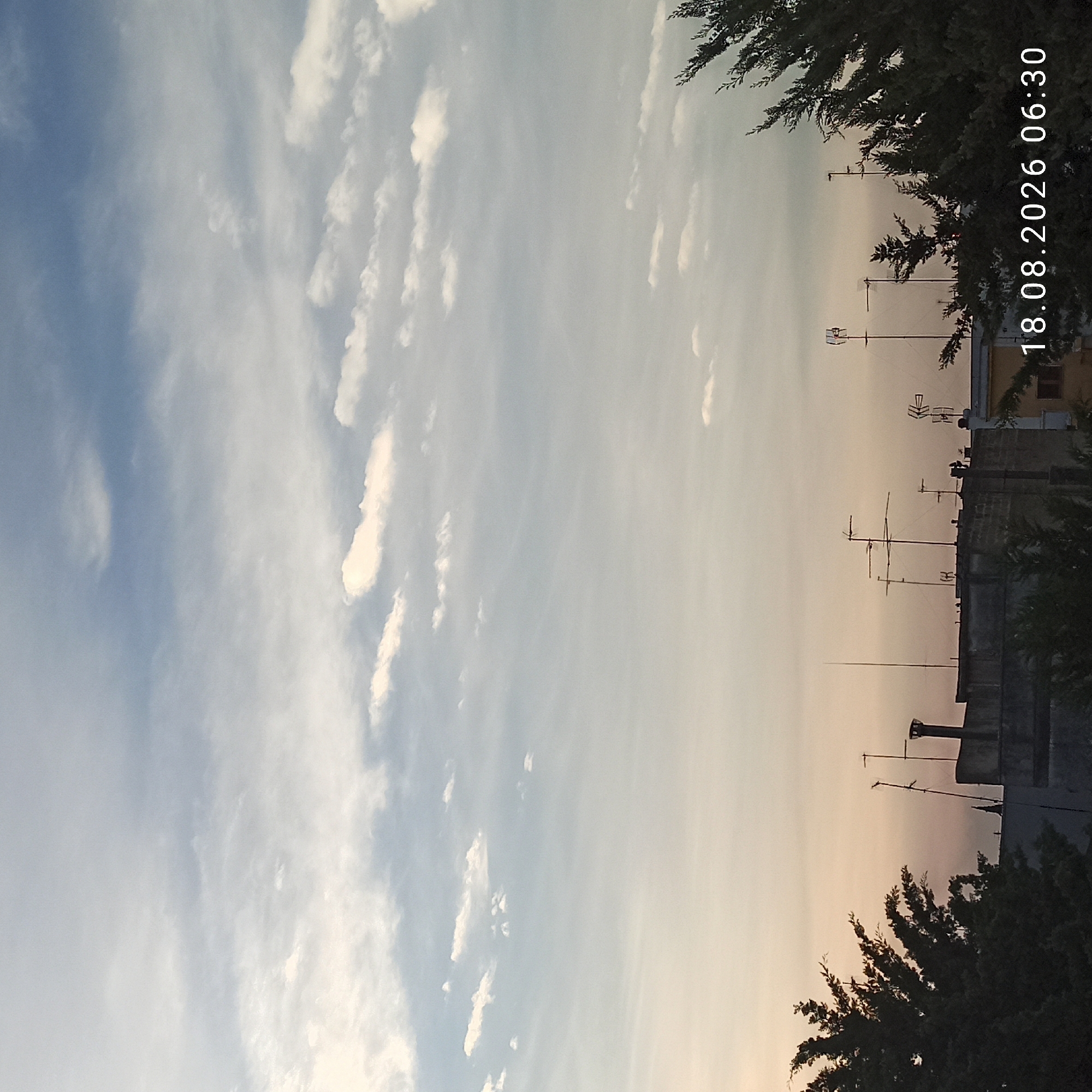 The image shows a cityscape with a sky full of clouds at dawn or dusk. The buildings in the foreground have several antennas on their roofs, which is typical for older buildings in many cities. There are also trees on the sides of the image. The date and time stamp in the lower right corner indicates that this image was taken on August 18, 2026, at 06:30. The warm tones in the sky suggest that the sun is either rising or setting. The presence of antennas and the architectural style of the buildings suggest that this photo was taken in an urban or suburban area. Bitonto, Italy, is known for its historical center with churches and older buildings, as well as a surrounding agricultural landscape. The image is consistent with a view from a rooftop in Bitonto, showing a mix of residential buildings and trees against a cloudy sky. There are no people visible in the image. The scene is quiet and still, capturing a moment of transition between day and night.