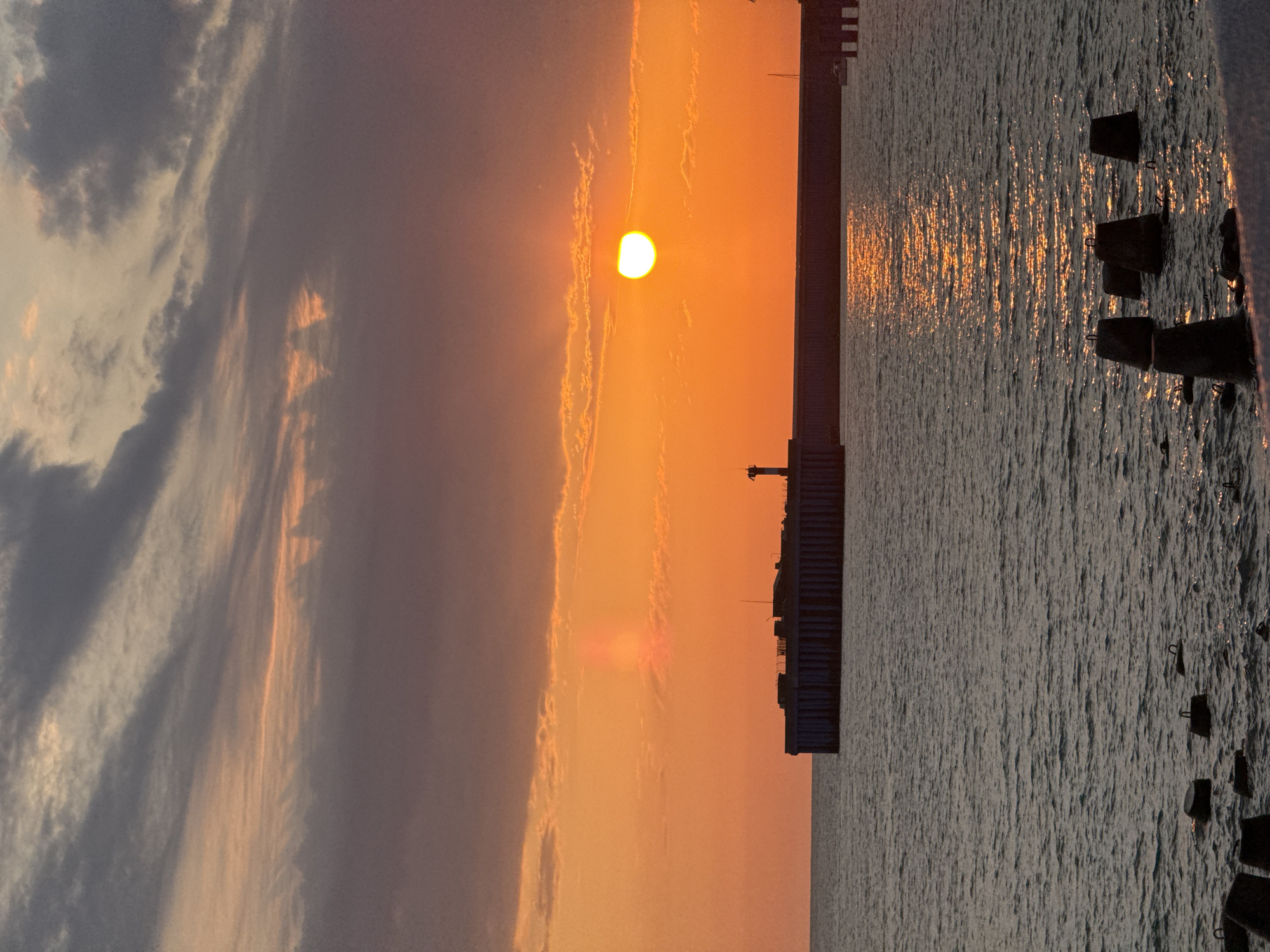 A sunset over the Black Sea in Sirius, Russia, is captured in this vertical, medium shot. The sun, a bright orb partially obscured by clouds, casts an orange glow across the sky, painting it with hues of orange, pink, and purple. The clouds, dark and wispy, are illuminated from behind by the sun, creating dramatic shadows and highlights.
In the foreground, the dark, rippling water of the Black Sea reflects the fiery sunset, creating a shimmering pathway of light. Scattered across the water are several concrete blocks, remnants of a breakwater or pier construction. In the mid-ground, a long, imposing breakwater or pier juts out into the sea from the left side of the frame. It is constructed of dark, rectangular concrete pillars and a flat top surface. A small lighthouse, a slender white structure with a dark top, stands on the breakwater.
The overall atmosphere is one of tranquil beauty, with the natural elements of the sea and sky dominating the scene. No people are visible in the image.