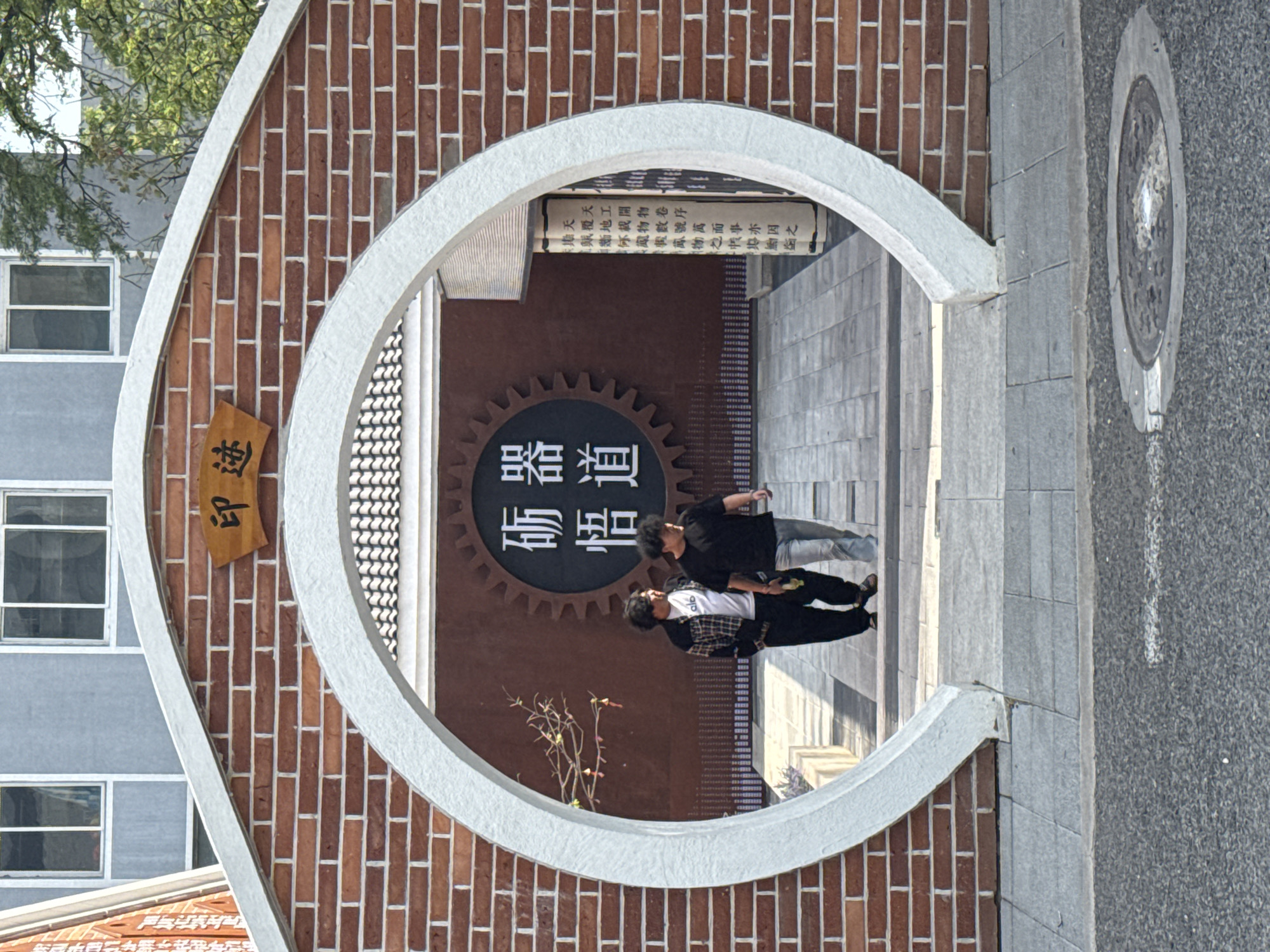 Two people are positioned within a circular opening of a brick structure. One person is wearing a black shirt and the other is wearing a white shirt under a plaid jacket. The opening is part of a decorative brick facade that also incorporates a sign in Chinese characters that reads "印迹" and another sign with the characters "砺悟" set within a gear symbol, indicating a focus on knowledge or experience. Above them is an inscription: "天工開物卷序天覆地載物數號萬而事亦因之"
The structure appears to be situated along a pedestrian area, evidenced by the paving and what appears to be a drain cover in the foreground. This location is in the city of Zhongtun, China.