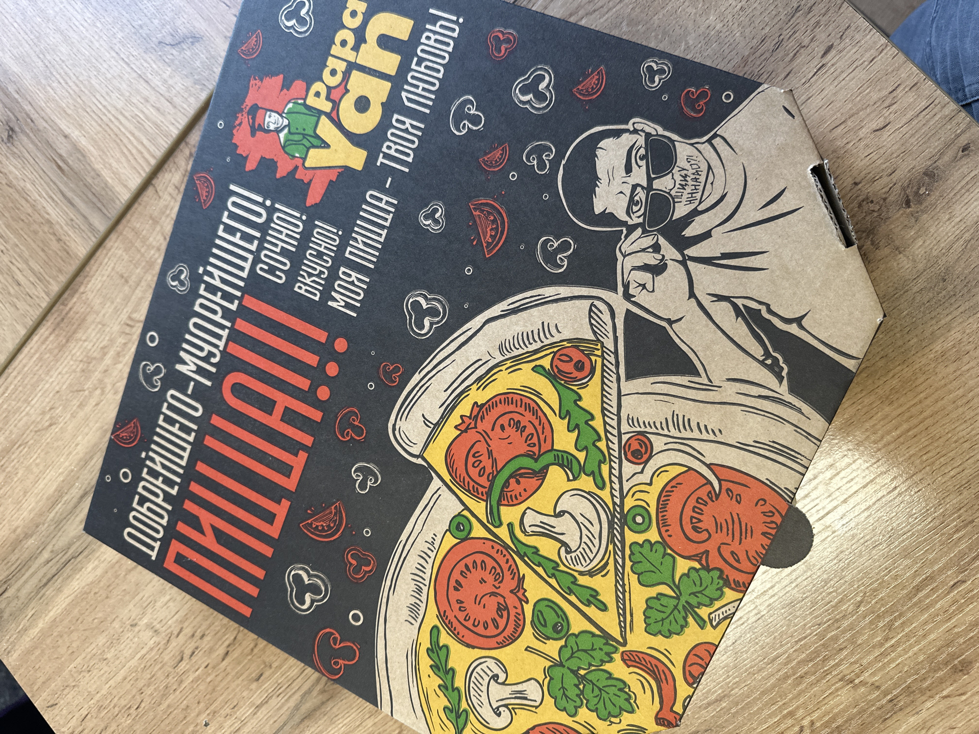 The image shows a pizza box with cartoon-style graphics. The box is dark in color with illustrations of pizza slices, mushrooms, peppers, and a man with sunglasses, likely representing the "Papa" from the brand "Papa Yan," printed on the box. Text in Russian is visible, including phrases that translate to "the kindest-wisest!", "pizza!!!", "tasty!", "my pizza - your love!". The pizza box is sitting on a wooden surface. The image suggests this is a pizza delivery or takeaway box from Papa Yan, possibly in Biysk, Russia, given the Russian text.