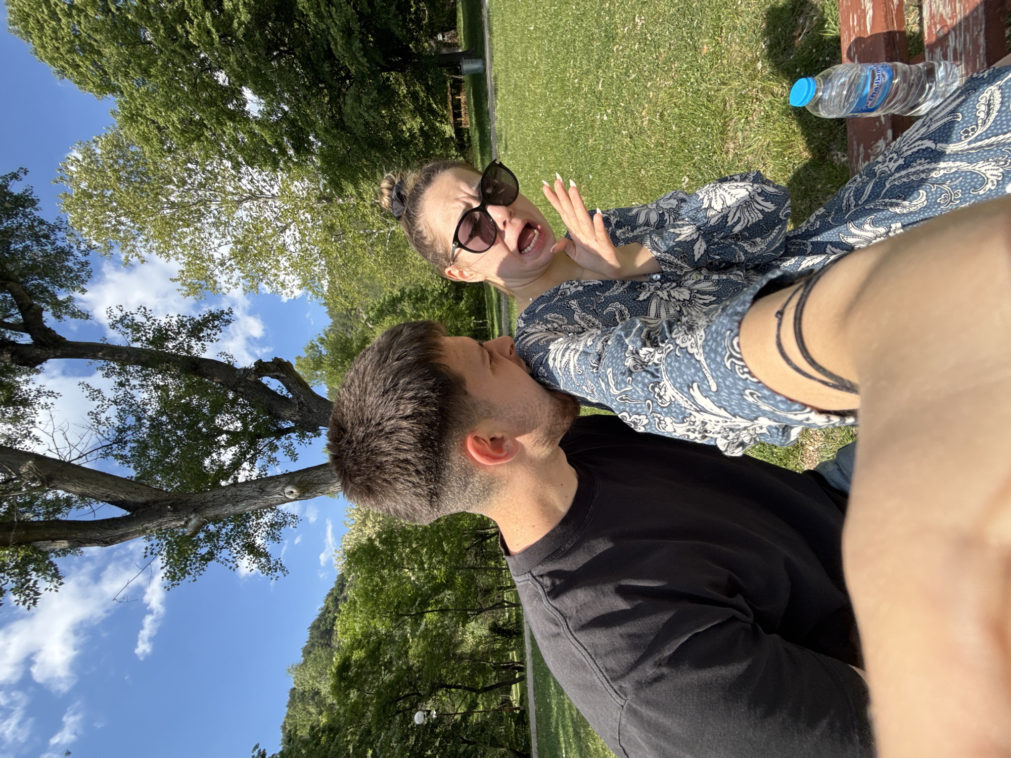 The image shows a man and a woman sitting on a bench in a park in Blagoevgrad, Bulgaria. The woman has dark hair pulled back, sunglasses, and is wearing a blue and white patterned dress. She is making a face with her mouth open and her hands raised. The man has short dark hair and is wearing a black t-shirt. He appears to be kissing or biting the woman's shoulder. A bottle of water sits on the bench next to the woman. Trees and grass are visible in the background, with a blue sky and some clouds. The photograph is taken at an angle, possibly by one of the individuals in the image.