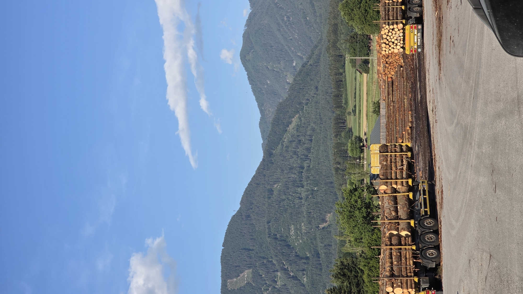 The image depicts an outdoor scene in Feistritz an der Drau, Austria, featuring a lumber yard. In the foreground, a large truck loaded with logs is visible, positioned on a paved surface. To the right of this truck, another truck also carrying logs is parked, with the text "matik" displayed on its rear. Extensive stacks of logs are arranged behind the trucks, forming a significant portion of the yard's inventory. The background is dominated by a verdant, tree-covered mountain range under a clear blue sky dotted with white clouds. Agricultural fields and scattered buildings are visible at the base of the mountains, suggesting a rural setting. The overall atmosphere is one of industrial activity related to timber processing, set against a backdrop of natural beauty.