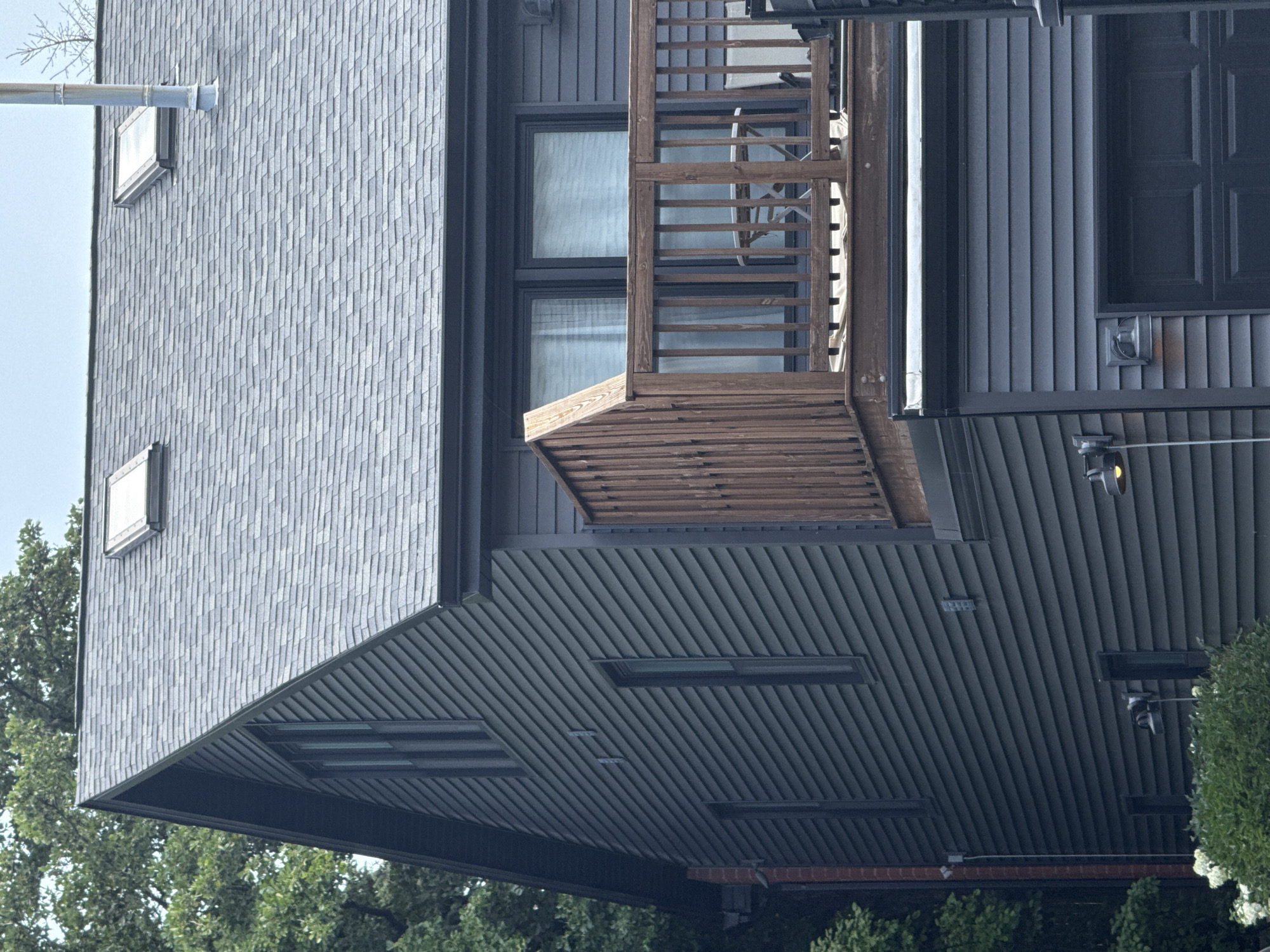 The image shows the exterior of a dark gray, two-story house with a gray asphalt shingle roof. There are two skylights on the roof and a metal chimney pipe. The house has dark gray siding with horizontal lap joints. On the second floor, there is a wooden deck with a railing. The deck has a small table and chairs visible on it. Below the deck, on the first floor, is a dark gray garage door. There are two outdoor lights mounted on the side of the house. The house is surrounded by lush green trees. No people are visible in the image. The scene appears to be a residential property in an urban or suburban setting, likely in Chicago, United States of America, given the architectural style and typical residential landscaping.