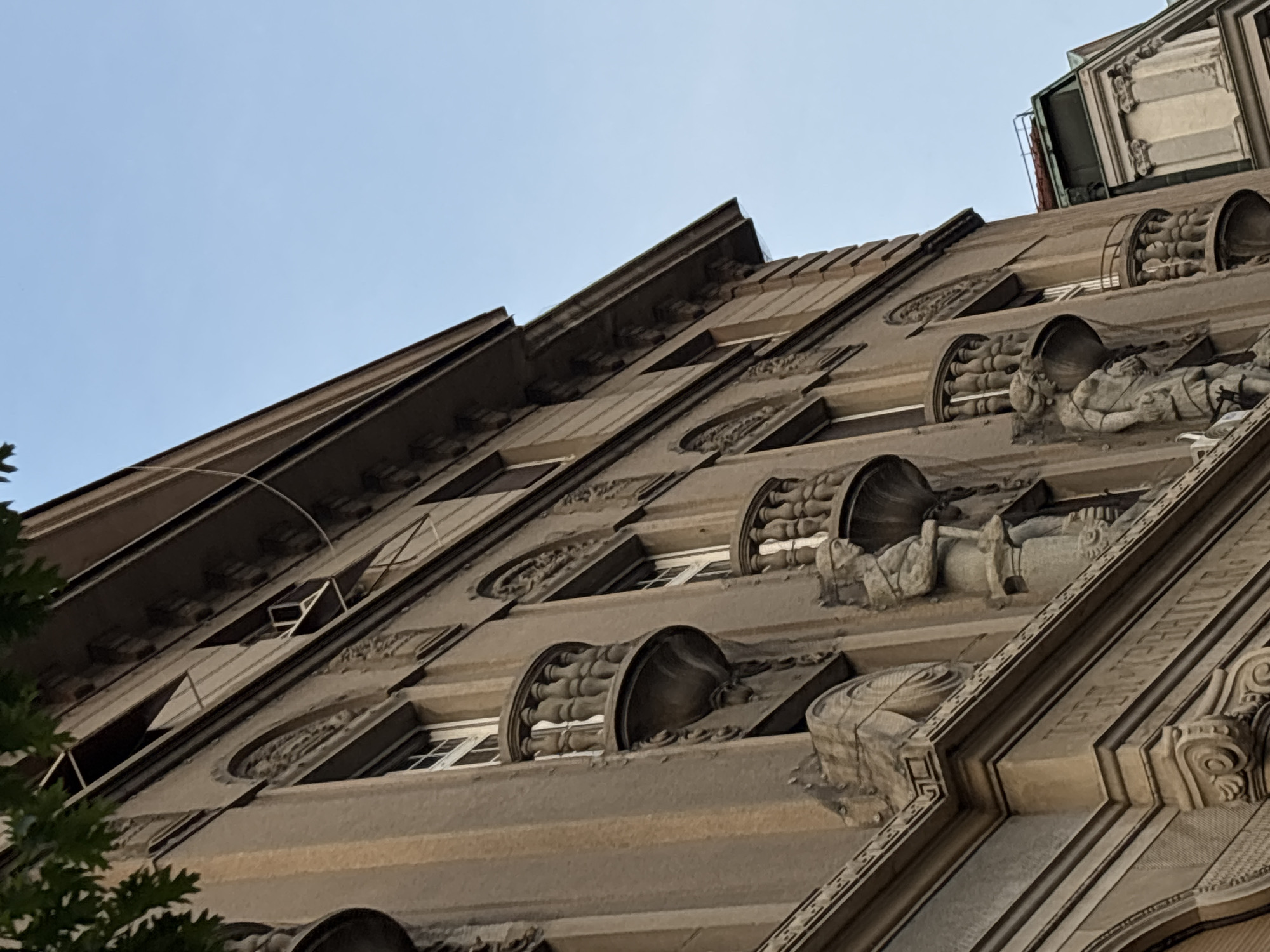 A low-angle, upward-tilted shot captures the facade of a building in Belgrade, Serbia. The building's architectural style is ornate, featuring intricate stone carvings, including relief sculptures of figures and decorative elements. The exterior is primarily a light brown or beige color. The windows are recessed and rectangular with visible panes and frames. The sky is pale blue and mostly clear. The building appears to be multi-storied. Some greenery is visible in the bottom left corner, potentially trees or foliage. The perspective of the shot emphasizes the height and grandeur of the building's facade.