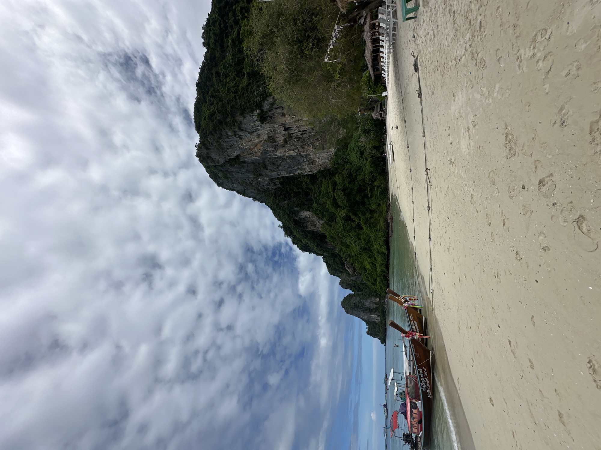 This is a photograph of a tropical beach scene in Ao Nang, Thailand. The foreground is dominated by a light-colored sandy beach with scattered footprints. In the mid-ground, several traditional longtail boats are moored near the shore. The water is calm and a light turquoise color. In the background, a large, steep, jungle-covered limestone cliff rises dramatically. The sky is partly cloudy with large white and gray clouds. Further back, more rocky islands covered in vegetation can be seen in the distance. On the beach, to the right of the frame, there are rows of empty lounge chairs and a small thatched-roof hut, suggesting a tourist resort. A few people are visible on the boats and in the shallow water. The overall impression is a serene and beautiful coastal landscape.