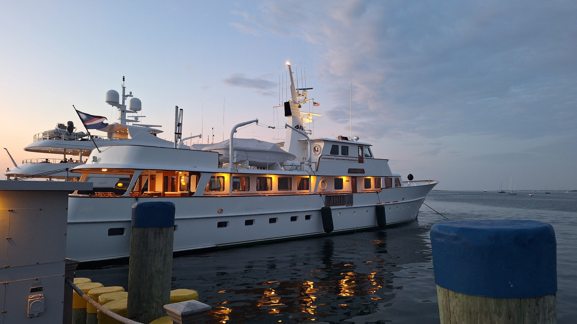 The image shows a large, white motor yacht docked in the city of Nantucket, United States of America. The yacht, named "OBELISQUE," is prominently featured, with its interior lights illuminated, creating a warm glow. The vessel is moored alongside a dock, with wooden pilings visible in the foreground, some capped with blue and yellow protective covers. A smaller, white yacht is also partially visible in the background, suggesting a marina or harbor setting. The sky is a dusky blue, with hints of a receding sunset, and scattered clouds. The water is calm, reflecting the lights of the yacht. There are no people readily discernible in the image, but the scene implies a quiet evening ambiance at the harbor.