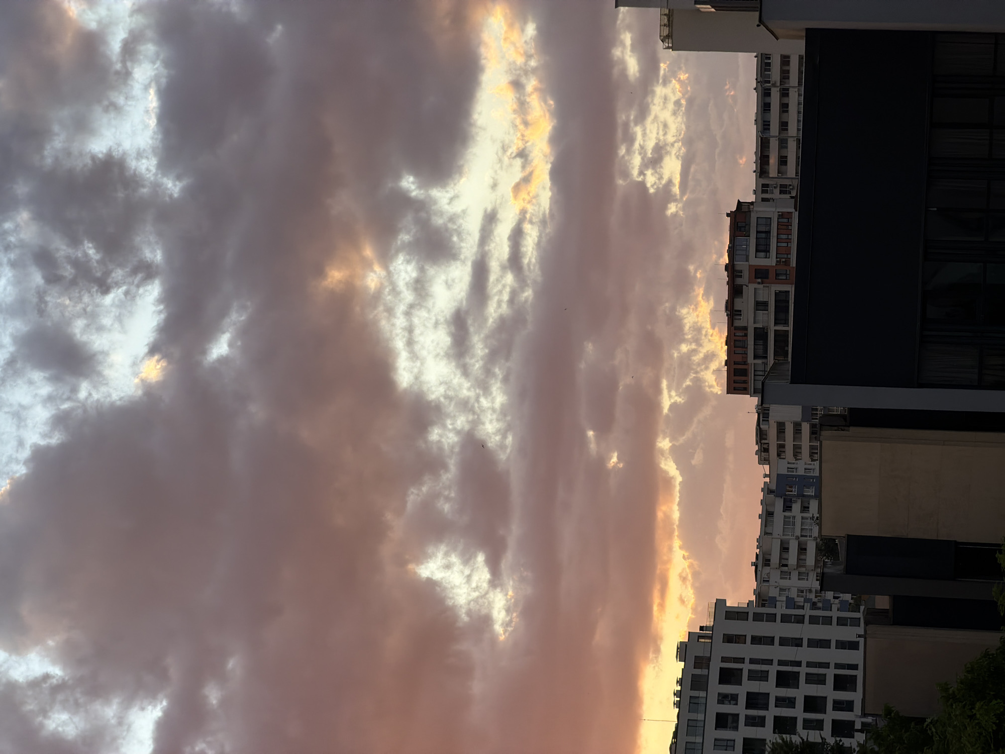 This image depicts a sunset scene over a city, likely Sofia, Bulgaria. The sky is filled with dramatic clouds painted in shades of pink, orange, and yellow, illuminated by the setting sun. Below the vibrant sky, a dense cluster of apartment buildings and other structures form the cityscape. The architecture appears to be modern, with multi-story residential buildings featuring numerous windows and balconies. Some buildings are white, while others are cream or have colorful facades. In the foreground, darker, more contemporary buildings with dark facades and visible windows are present. The overall impression is one of an urban environment bathed in the warm, fading light of dusk. There are no discernible people or specific actions captured in this still image.