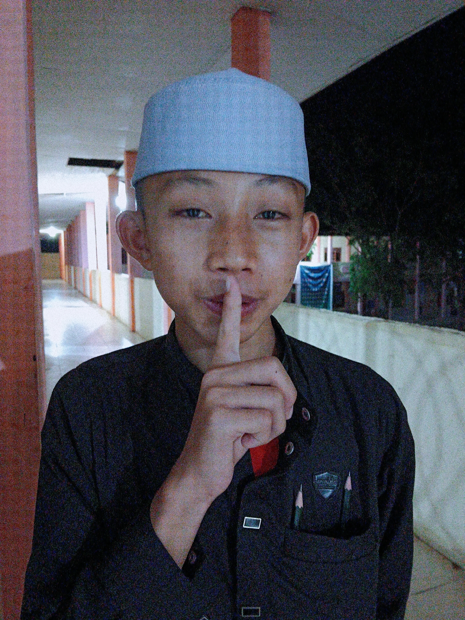A young person wearing a light blue cap and a dark button-up shirt stands indoors, holding a finger to their lips in a gesture for silence. The shirt has a pocket with two pencils sticking out. The background appears to be a hallway with light-colored walls and a darker area further in. The location is Lhokseumawe, Indonesia.