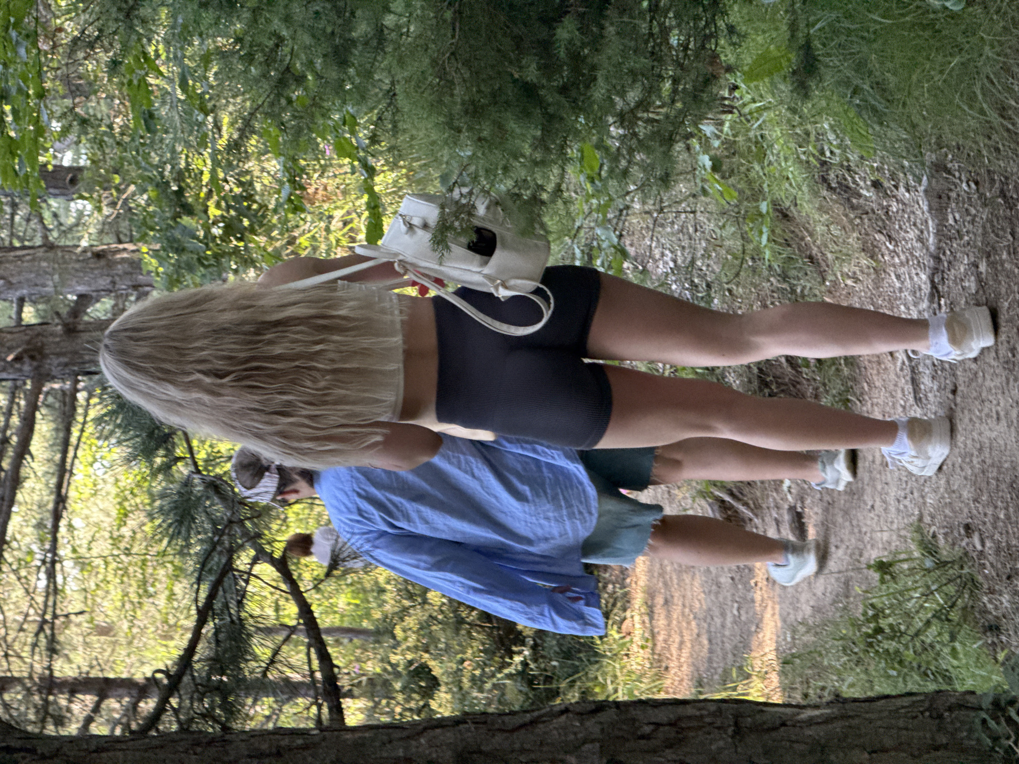 Two people are walking away from the camera on a dirt path through a wooded area. The person in the foreground is a blonde female wearing a white crop top, dark grey athletic shorts, and white sneakers. She has a white backpack with a red strap detail slung over her right shoulder. The person in the background, to the left, appears to be wearing a light blue oversized button-down shirt and light blue shorts. They are also wearing a visor and light-colored athletic shoes. In the far background, another person with short reddish hair is visible. The environment consists of trees, dense green foliage, and dappled sunlight filtering through the branches. The path is dusty and appears well-trodden. The scene is outdoors. The location is not explicitly visible in the image, and Zaprudnoe, Ukraine cannot be confirmed from the visual information provided.