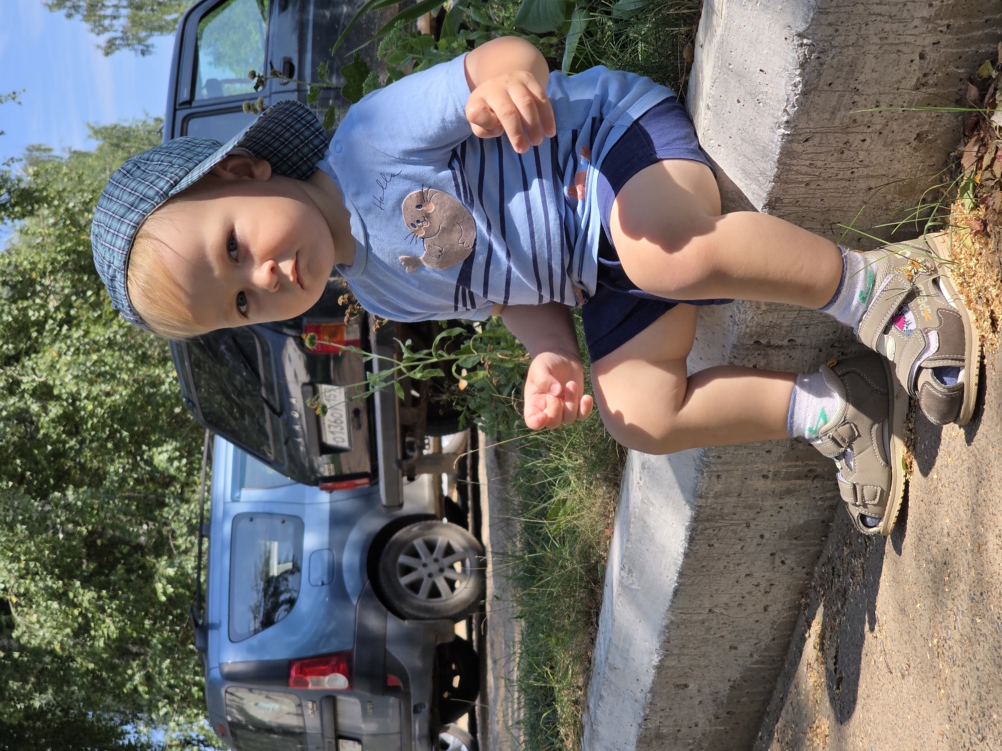 A fair-skinned, blonde-haired toddler wearing a blue and green plaid baseball cap and a light blue t-shirt with horizontal stripes and a seal graphic is sitting on a concrete curb. The child is looking directly at the camera with a neutral expression. The t-shirt has the word "Hello" written in cursive above the seal graphic. The toddler is also wearing dark blue shorts, white socks with green accents, and gray velcro sandals. In the background, two dark-colored SUVs are parked, with trees and foliage surrounding them. The image appears to have been taken outdoors on a sunny day. The license plate on the rear of the black SUV reads "0136OX159," which is a Russian license plate format, suggesting a location in Russia. Specifically, the "159" in the license plate corresponds to the Perm Krai region, where the city of Chaikovsky is located.