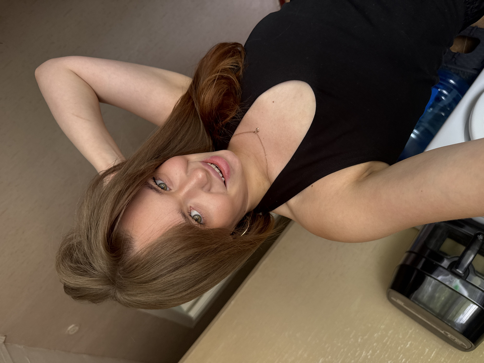 A young woman with light brown hair and green eyes smiles at the camera, holding one arm behind her head. She is wearing a black tank top and a thin gold necklace. The background appears to be indoors, with a beige wall and a light-colored ceiling. To the left of the woman, a black appliance with a silver trim is visible. To the right, several blue water bottles are stacked. The scene is likely a domestic setting in Chaikovsky, Russia.