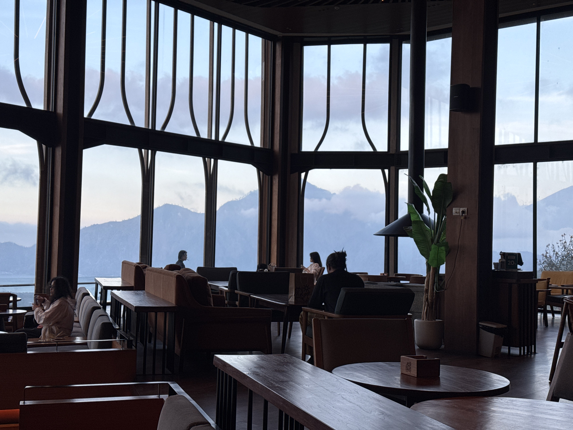 This image depicts the interior of a spacious cafe or restaurant located in Kintamani, Indonesia, with large windows offering a panoramic view of the surrounding mountains and a body of water. The architecture features high ceilings with exposed beams and a dark wood aesthetic.
Inside, several seating areas are visible, including plush sofas and individual chairs arranged around tables. A few patrons are present, engaged in their own activities. On the left, a woman in a light-colored jacket is seated on a sofa, holding a phone. Further back, another individual appears to be looking out the window. On the right side of the frame, a person with their hair in a bun is seated at a table, facing away from the camera.
The environment outside is characterized by rolling hills or mountains under a cloudy sky, with hints of a large lake or sea in the distance. The lighting suggests it is either early morning or late afternoon, with soft, diffused light.
Notable objects within the cafe include a large potted plant, a fireplace suspended from the ceiling, and various tables and chairs. A trash can and a small stand with what appears to be a menu or sign are also visible. The overall atmosphere seems calm and relaxed.