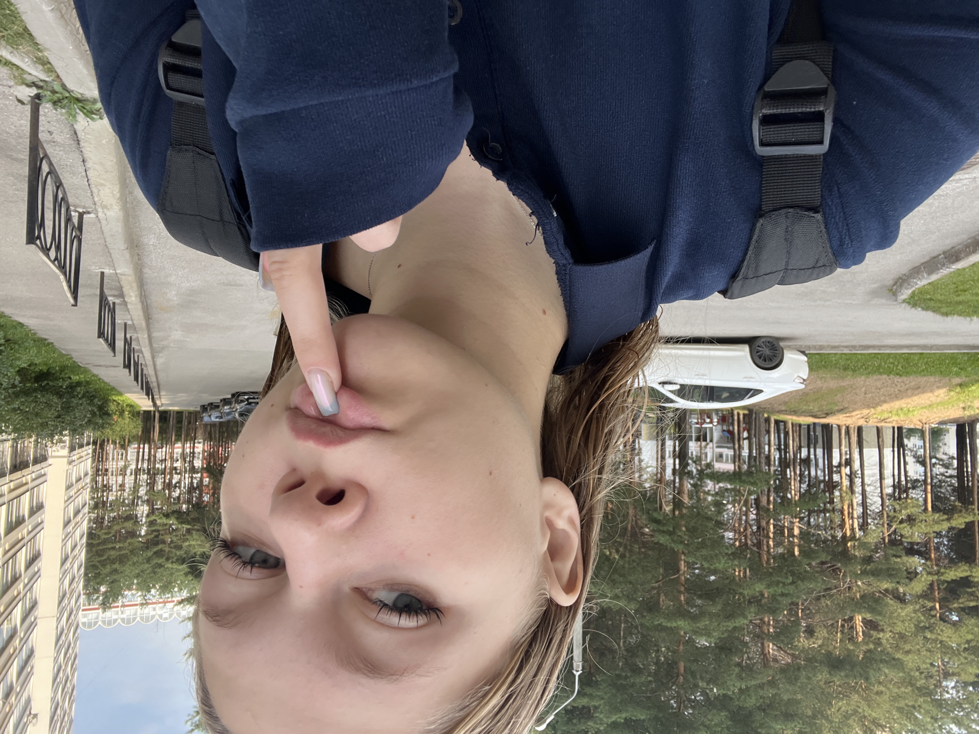 A young Caucasian woman with blonde hair stands outdoors, looking at the camera. She is wearing a dark blue collared shirt and a black backpack. Her right hand is raised, with her index finger touching her lips. In the background, there are tall pine trees, apartment buildings, parked cars, and a sidewalk. The setting appears to be an urban residential area.