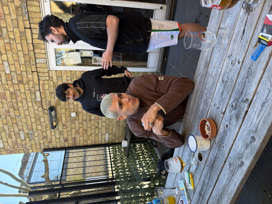 An outdoor pub garden in London, United Kingdom, is depicted under bright daylight. Three adult males are present. A man with bleached blonde hair, wearing a brown zipped Belstaff jacket, sits at a weathered wooden picnic table. He wears a prominent ring on his left hand, which is clasped on the table, and he looks to his left with a pensive expression. On the table in front of him are an empty glass, an open Marlboro cigarette pack with a cigarette, a half-full glass of lager, a white enamel mug with "TRAY" written on it, and a terracotta ashtray containing cigarette butts. Behind him, two men stand. The man on the left, with dark curly hair and a black hooded sweatshirt, holds a clear glass and smiles slightly, looking right. The man on the right, with dark hair and a beard, wears a black short-sleeved top over a black gilet and white New Balance shorts with green and orange stripes. He holds a clear glass and looks left, engaging with the man in the hoodie. On the table to the right, an empty beer glass, a partially visible glass of lager, and Rizla rolling papers are present. The background features a light-colored brick wall with a mounted heater, a white-framed glass door, and a black metal fence with artificial foliage, beyond which a building with a large glass window is visible.