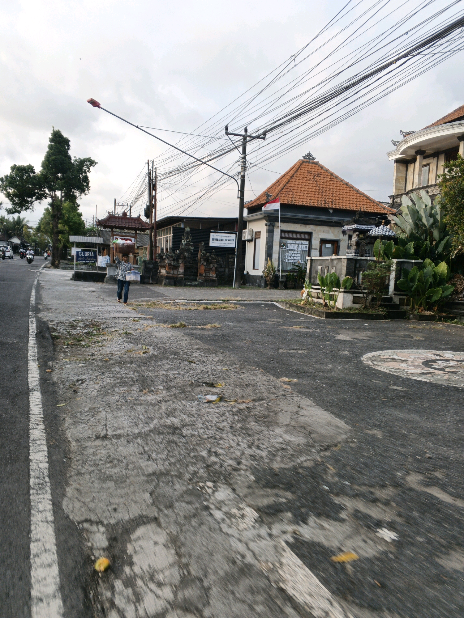 A street scene in Sukawati, Indonesia. A person is walking on the right side of the road, carrying a plastic container and a green object. The road is paved and appears to be in moderate condition, with some cracks and debris. There are several buildings on the right side of the road, including one with a red tiled roof and a Balinese-style shrine in front. A sign with the text "LUMBUNG DEWATA" is visible on one of the buildings. The street is lined with trees and utility poles with numerous electrical wires. The sky is overcast. In the background, a few motorcycles are parked.