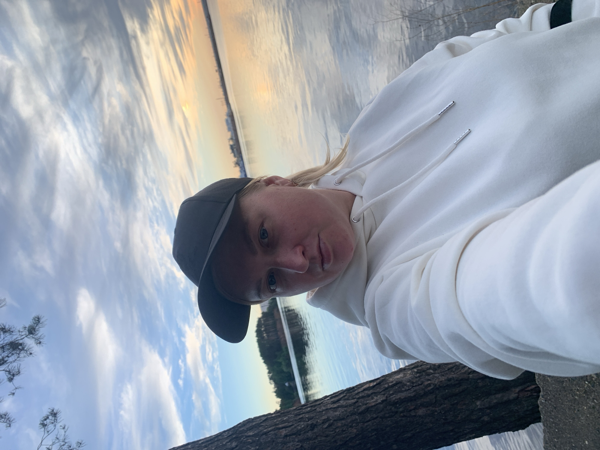 A person wearing a white hoodie and a black baseball cap is positioned on a tree trunk, likely near a body of water at sunset. The scene includes a shoreline or distant buildings across the water. The picture was taken outside in what looks to be Chaikovsky, Russia, but there is no location metadata available to confirm.