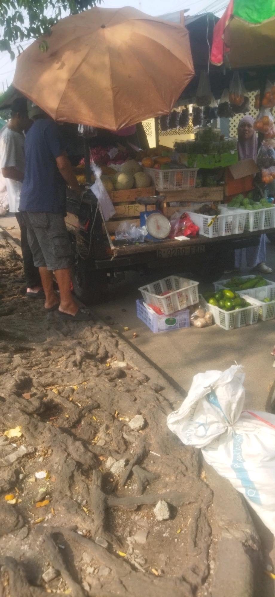 The image depicts an outdoor scene at a street market in Depok, Indonesia. A fruit and vegetable stall is set up on the back of a small truck, which is partially visible. The stall is covered by a large, tan umbrella, providing shade. Various fruits and vegetables are displayed in white plastic baskets, including what appear to be avocados, cucumbers, and possibly melons. Some bags of produce hang in the background. A traditional scale is placed on the truck bed. Two individuals, both men, are standing near the stall. One man, wearing a blue t-shirt and grey shorts, is facing away from the camera. Another man, wearing a white t-shirt, is partially visible to the left. A woman wearing a light purple hijab is also visible in the background, near the stall. The ground in the foreground is uneven and appears to be dirt with exposed roots and some scattered debris, suggesting a somewhat unpaved or weathered area typical of local markets. A large white sack, possibly containing goods, is prominently placed in the lower right portion of the frame.