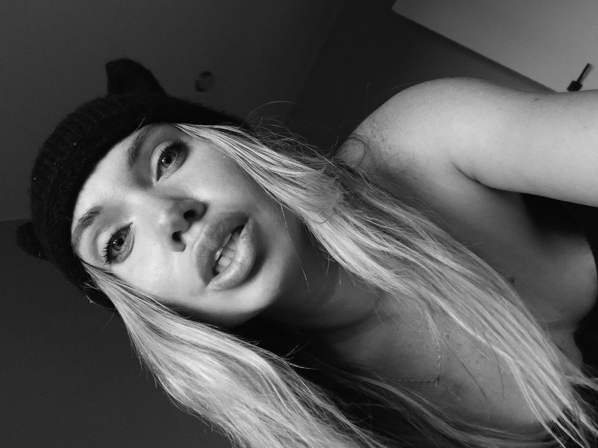 A black and white, close-up, high-angle shot depicts a young woman with light-colored, long hair. She is wearing a dark, knitted beanie with small, pointed extensions resembling cat ears. The woman's face is angled towards the viewer, showcasing her prominent lips. Her gaze is direct. The image's background is partially visible, with a wall and a lighting fixture vaguely discernable. The image provides no information to associate it with Votkinsk, Russia, nor is there anything identifiable in the picture pointing to a particular location.