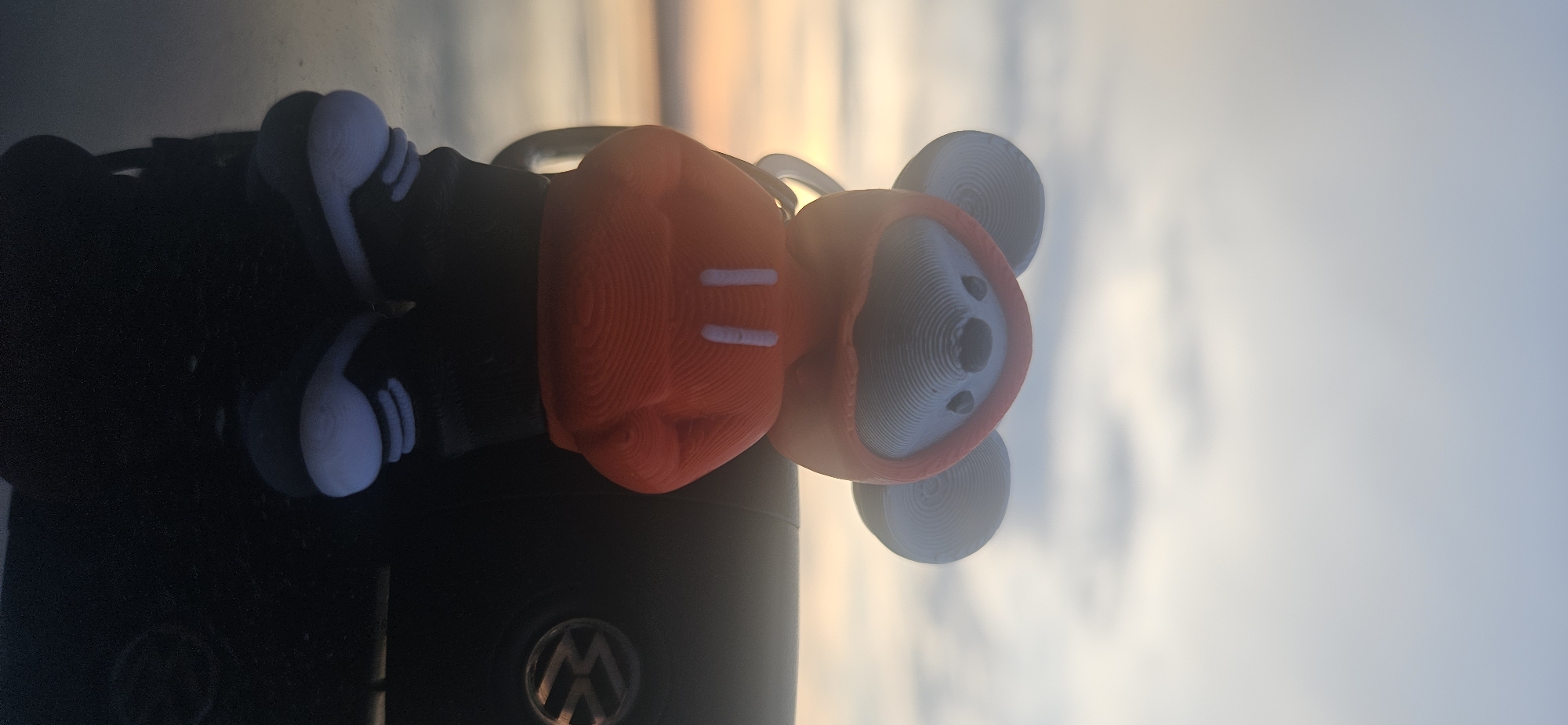A 3D printed figurine of a mouse wearing a red hoodie and black pants stands on a dark, reflective surface, possibly a car. The mouse has large, round ears and wears white sneakers with black laces. Behind the figurine, to the right, is the dark exterior of a car, featuring a visible Volkswagen logo. The background consists of a hazy sky with streaks of orange and yellow, indicating either a sunset or sunrise, with a body of water visible in the distance. The overall lighting is soft and diffused. The location of Momin Prohod, Bulgaria is not discernible from the image.