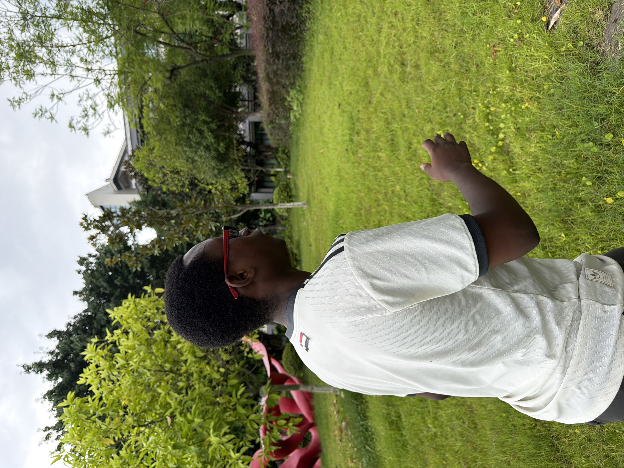 In the city of Jiangjin, China, a young Black person wearing a white and black Adidas jersey and red-framed glasses stands in a grassy area. The individual is facing to the right, with their left arm slightly raised and hand open, as if interacting with something unseen or gesturing. Behind them, lush green trees and bushes form a natural backdrop, with a modern building visible in the distance. The sky is overcast, suggesting a cloudy day. A large, abstract red sculpture is partially visible on the left side of the frame, adding a touch of artistic interest to the outdoor scene. The ground is covered in bright green grass, with scattered small yellow flowers.