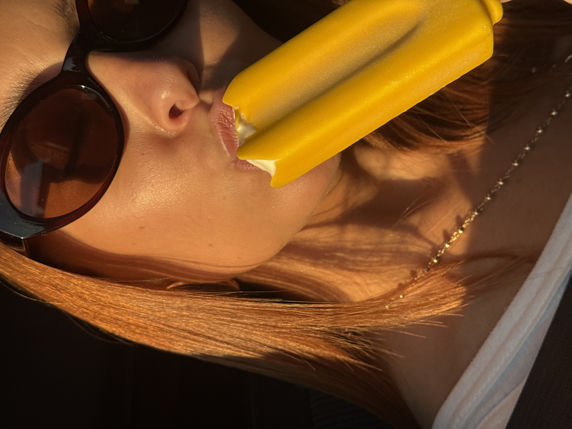 A close-up, waist-up shot captures a person with reddish-orange hair and light skin, wearing dark sunglasses and a white top, eating a yellow popsicle. The person is biting into the popsicle, which has a white interior. The scene is illuminated by golden hour sunlight, creating strong shadows and highlights on the person's face and hair. A delicate gold necklace with small beads is visible around the person's neck. The background is dark and out of focus. There is no identifiable location in the city of Chaikovsky, Russia, visible in this image.