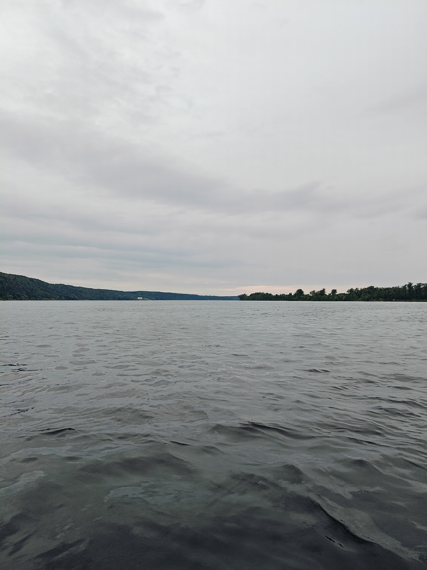 The image depicts a serene, wide body of water under a cloudy sky. The water exhibits gentle ripples, reflecting the overcast conditions. In the distance, the shoreline is visible, lined with lush green trees and a gentle slope on the left side of the frame. A few sparse structures can be seen along the distant coastline, one appearing as a white building with a distinct rectangular shape. The scene evokes a sense of calm and tranquility, suggesting it might be a quiet, natural setting, possibly a lake or a wide river. The overall impression is one of a peaceful, overcast day in a natural environment. The location context of Olkhovka, Russia, further reinforces the likelihood of a natural, possibly rural or semi-rural landscape. There is no visible text in the image.