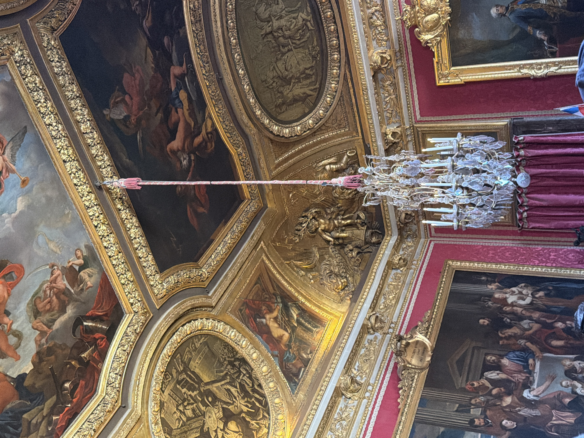 This is a photograph of the interior of a room in the Palace of Versailles, France. The room features ornate gilded ceilings with large paintings depicting mythological or historical scenes. A grand crystal chandelier hangs from the ceiling, illuminated by several candles. The walls are adorned with large framed paintings, including a scene of what appears to be a biblical feast on the left and a portrait of a man in regal attire on the right. The walls themselves are covered in a rich red fabric, possibly damask, with gilded decorative moldings running along the top. Red velvet curtains are visible, framing the paintings and adding to the opulent decor. The overall impression is one of extreme luxury and grandeur characteristic of the Palace of Versailles.
