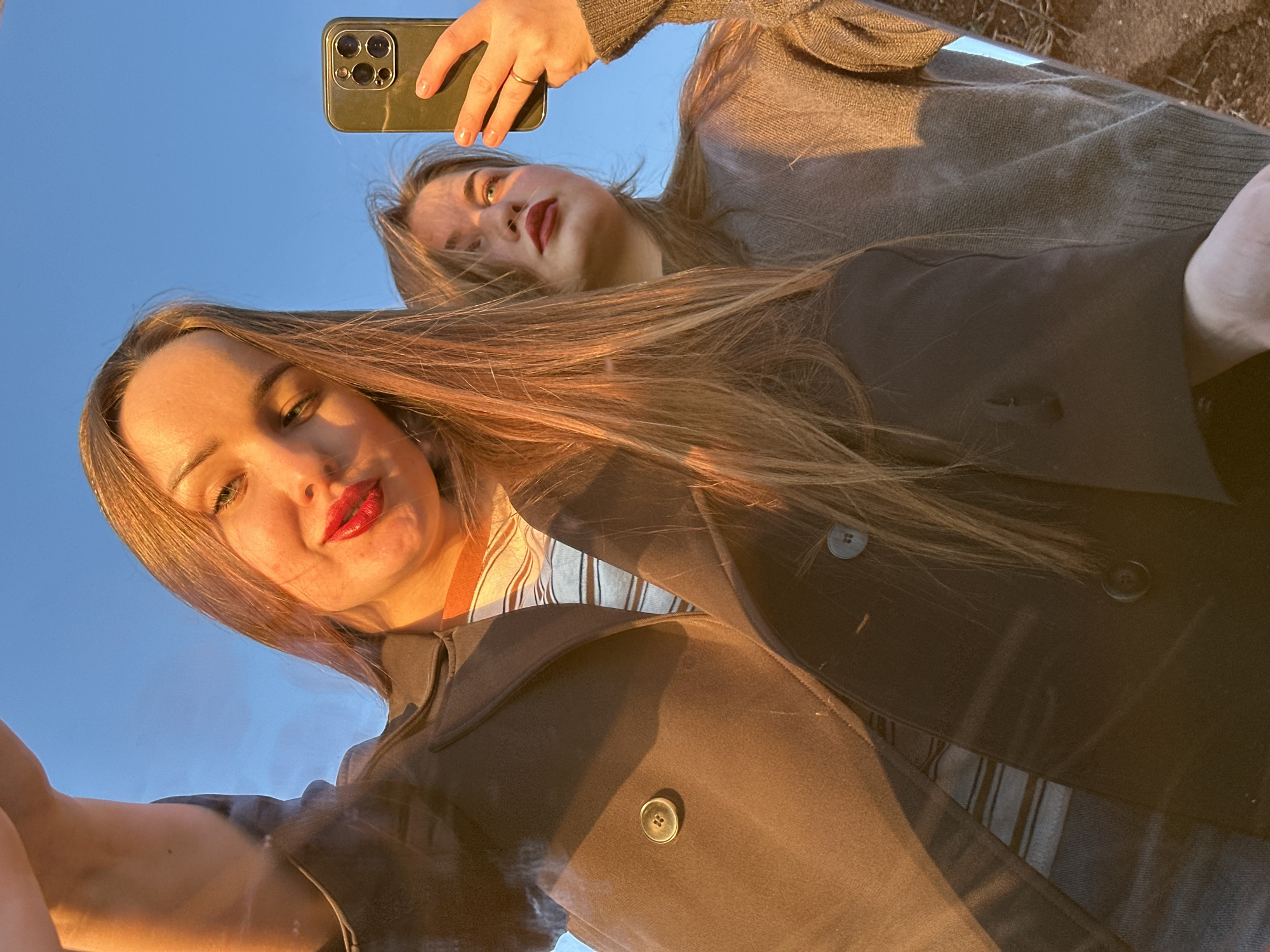 Two women are taking a selfie in a reflective surface. The woman in the foreground is wearing a black coat and has red lipstick. The other woman, partially visible in the reflection above, is wearing a brown sweater and holding a phone. The background appears to be a clear, blue sky. The setting is likely outdoors, possibly in Ольховка, Russia.