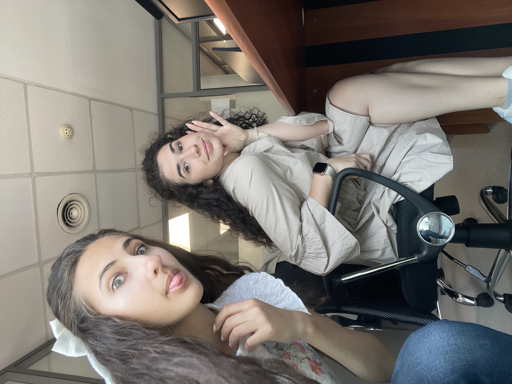 Two young women are seated at desks in what appears to be an office environment. The woman on the left, with long dark hair adorned with a white bow, is closer to the camera and makes a playful gesture with her tongue out. She is wearing a white top with floral print. The woman on the right, with curly dark hair, is wearing a striped dress and rests her chin on her hand, smiling. Both women are seated in office chairs. The background includes office furniture, a glass partition, and ceiling tiles with lighting fixtures and ventilation. The image was taken indoors, likely in an office building. The mention of Sofia, Bulgaria, is not visually supported by the provided image content.