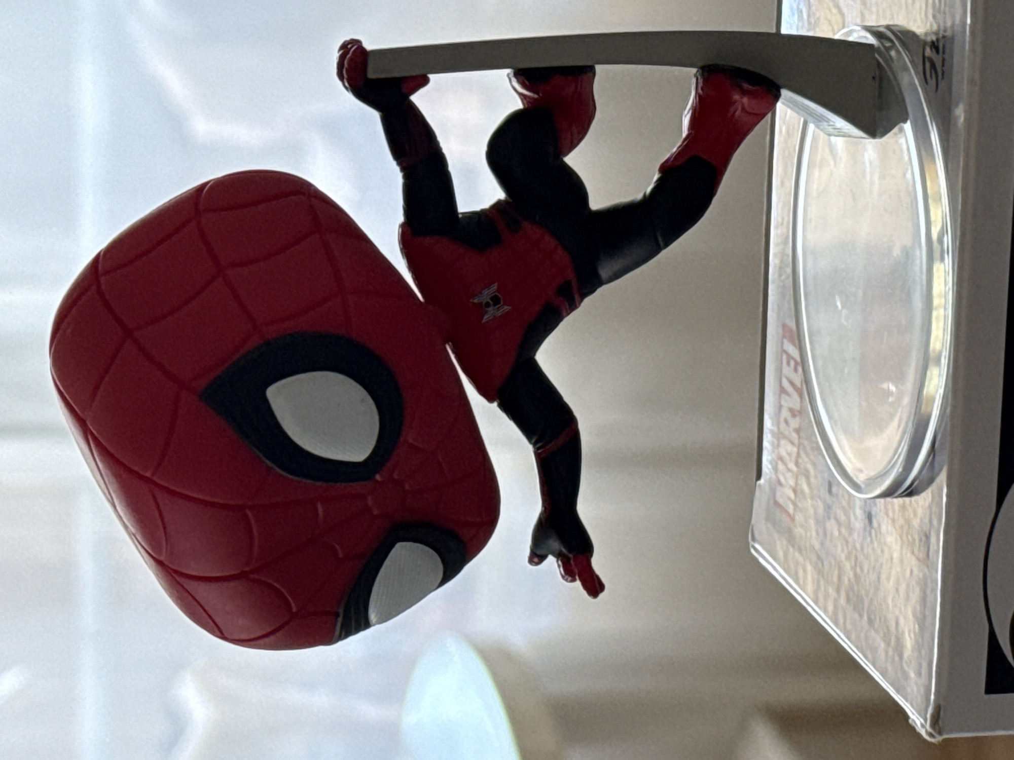 A close-up, eye-level shot shows a Spiderman Funko Pop figure in motion. The figure is red and black, with Spiderman's signature web pattern on his mask and suit. He is posed as if mid-swing, with one hand outstretched and the other gripping a silver pole. The figure is attached to a clear plastic base, which rests on top of a Marvel-themed box. The background is out of focus, appearing as a softly lit, neutral wall. The lighting creates a slight silhouette effect on the figure. The image is sharp and detailed, focusing on the Spiderman figure and its immediate surroundings. There is no indication of the city of Bad Sachsa, Germany in this image.