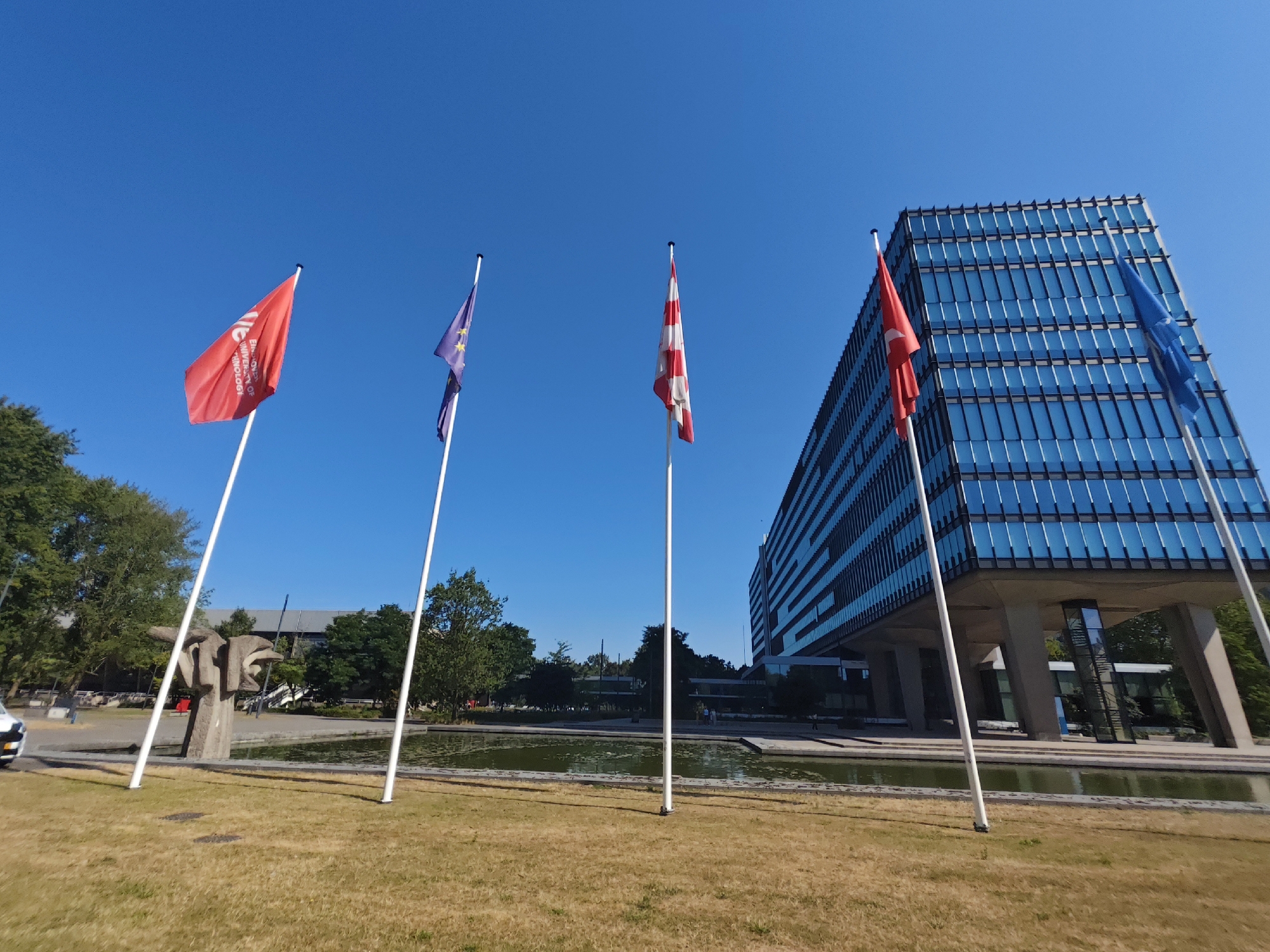 The image shows a wide-angle view of the TU/e park at the Eindhoven University of Technology in Eindhoven, Netherlands. In the foreground, a well-maintained lawn is visible, with dry, light brown grass. To the left, a modern sculpture made of concrete stands near a body of water. Several flagpoles are positioned in the foreground and middle ground, with flags flying from them. From left to right, the flags are red with white text, the European Union flag, a red and white striped flag, the Turkish flag, and another blue flag, possibly the flag of the Netherlands. In the background, a multi-story building with a glass facade dominates the scene. The building is supported by large concrete pillars, and a pond or water feature is situated in front of it. Trees are visible on the left and in the background, and the sky is clear and blue. The overall impression is of a bright, sunny day at a university campus.