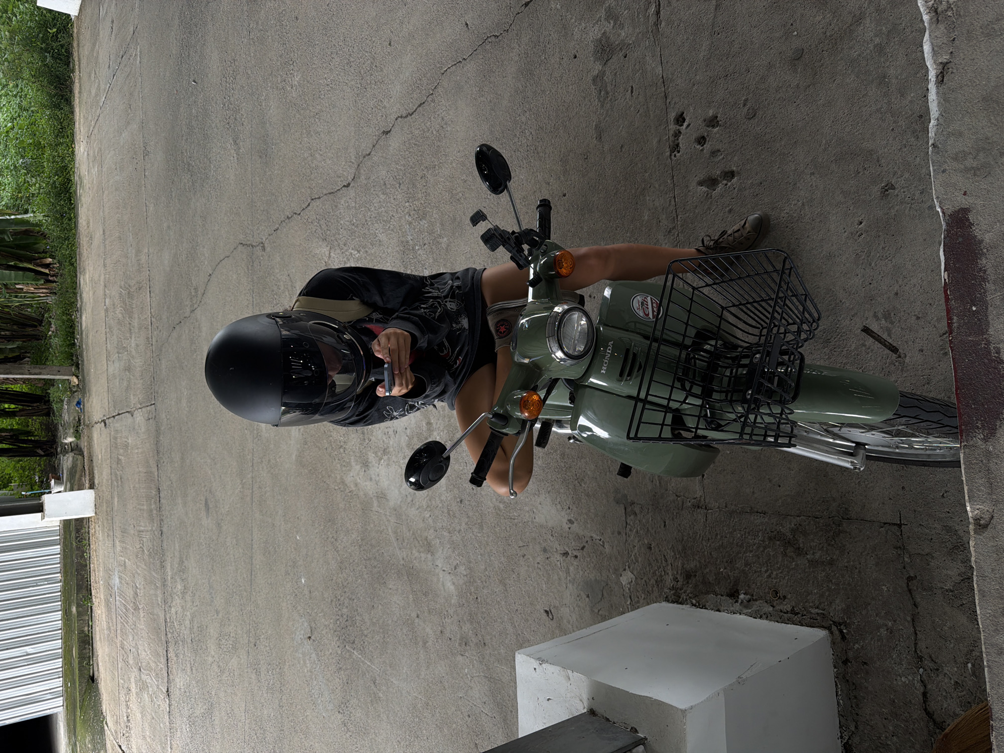 A person wearing a black helmet sits on a light green Honda motorcycle. The motorcycle has a black basket on the front. The person is wearing a dark-colored shirt and dark shorts, and brown shoes. They are holding a smartphone in their right hand. The motorcycle is parked on a concrete surface. To the left of the motorcycle, there is a section of corrugated metal siding and some greenery. To the right, there is a white concrete block. The scene appears to be outdoors, possibly in a driveway or parking area. The lighting suggests it is daytime. The image appears to be taken from a high angle, looking down at the subject and motorcycle. The concrete surface has visible cracks and textures. The overall atmosphere is casual and everyday. The word "HONDA" is visible on the side of the motorcycle.