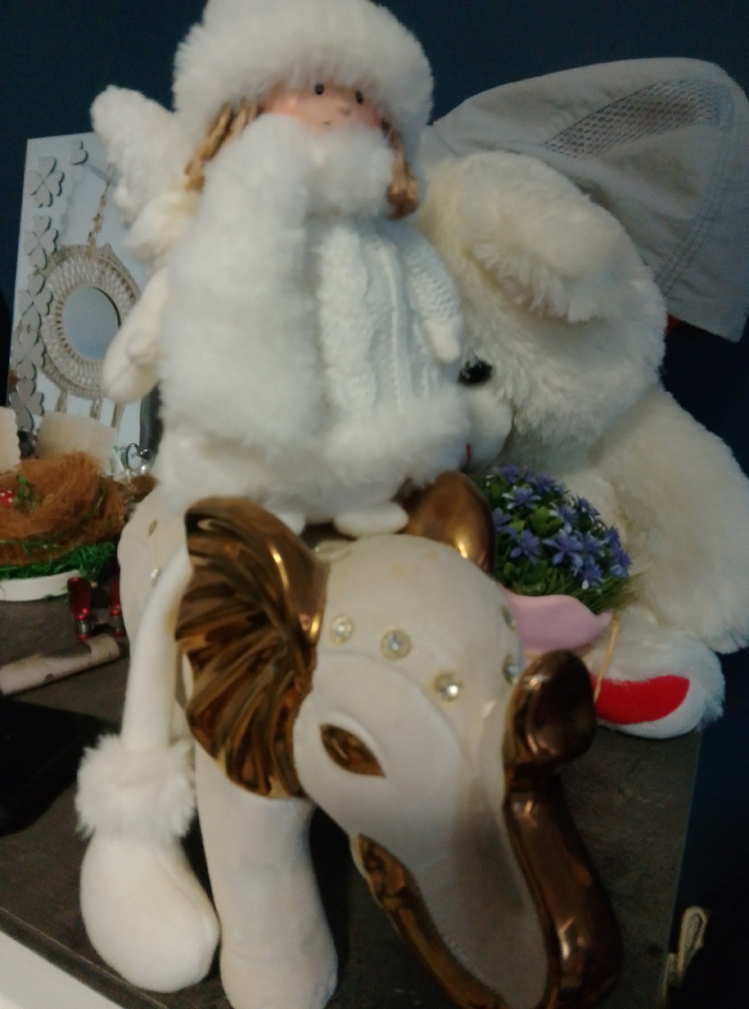 The image depicts a close-up of two stuffed toys arranged on a dark, flat surface, possibly a shelf or table. On the left is a white, plush angel figurine with feathered wings, a white knitted garment, and blonde hair. The angel is positioned atop a white, ceramic elephant. This elephant figurine is adorned with gold detailing on its tusks, ears, and legs, and has small, clear rhinestones embedded on its body. A cluster of small, artificial blue flowers in a pink holder is placed near the elephant's head. Behind the angel and elephant, a large white plush bear is visible, partially obscuring the angel's wing. The bear appears to be wearing a light gray cloth draped over its back. To the left of the angel, a decorative item resembling a woven circular frame with intricate patterns is visible, along with a small brown object that looks like a bird's nest with some green material inside. The background is dark, with hints of other objects suggesting a cluttered indoor environment. There is no indication of the city of Wedel, Germany, in the image itself.