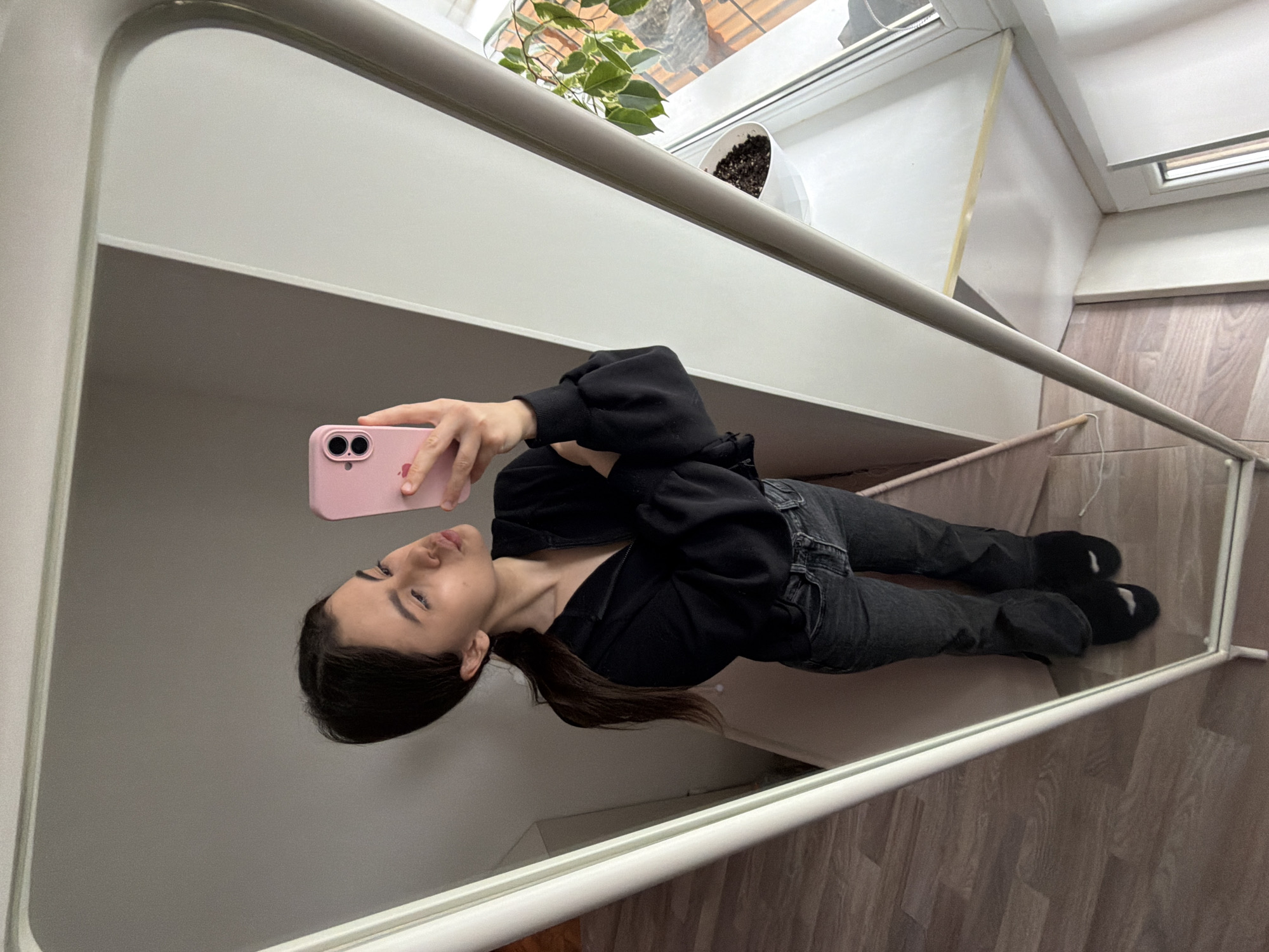 This image, taken in Chaikovsky, Russia, presents a mirror selfie of a person indoors. The individual, appearing to be a young woman, is reflected in a large, white-framed mirror, holding a pink smartphone (an iPhone, identifiable by its dual camera bumps) to capture the image. The photo is rotated 90 degrees counter-clockwise, making the person appear horizontal.
The subject is dressed casually in a black hooded top or jacket, dark grey jeans, and dark-colored socks with white accents on the heels. Their dark hair is pulled back, and they have a neutral to slightly relaxed expression while looking at their reflection.
The interior setting visible in the mirror's reflection includes a bright window with a plant in a white geometric pot on a windowsill or shelf. The floor is covered with light-toned wooden laminate. The walls are a clean white or light grey, and the space appears well-lit, suggesting daytime. No discernible text is visible in the image. The scene captures a simple, everyday moment in a tidy, modern-looking room.