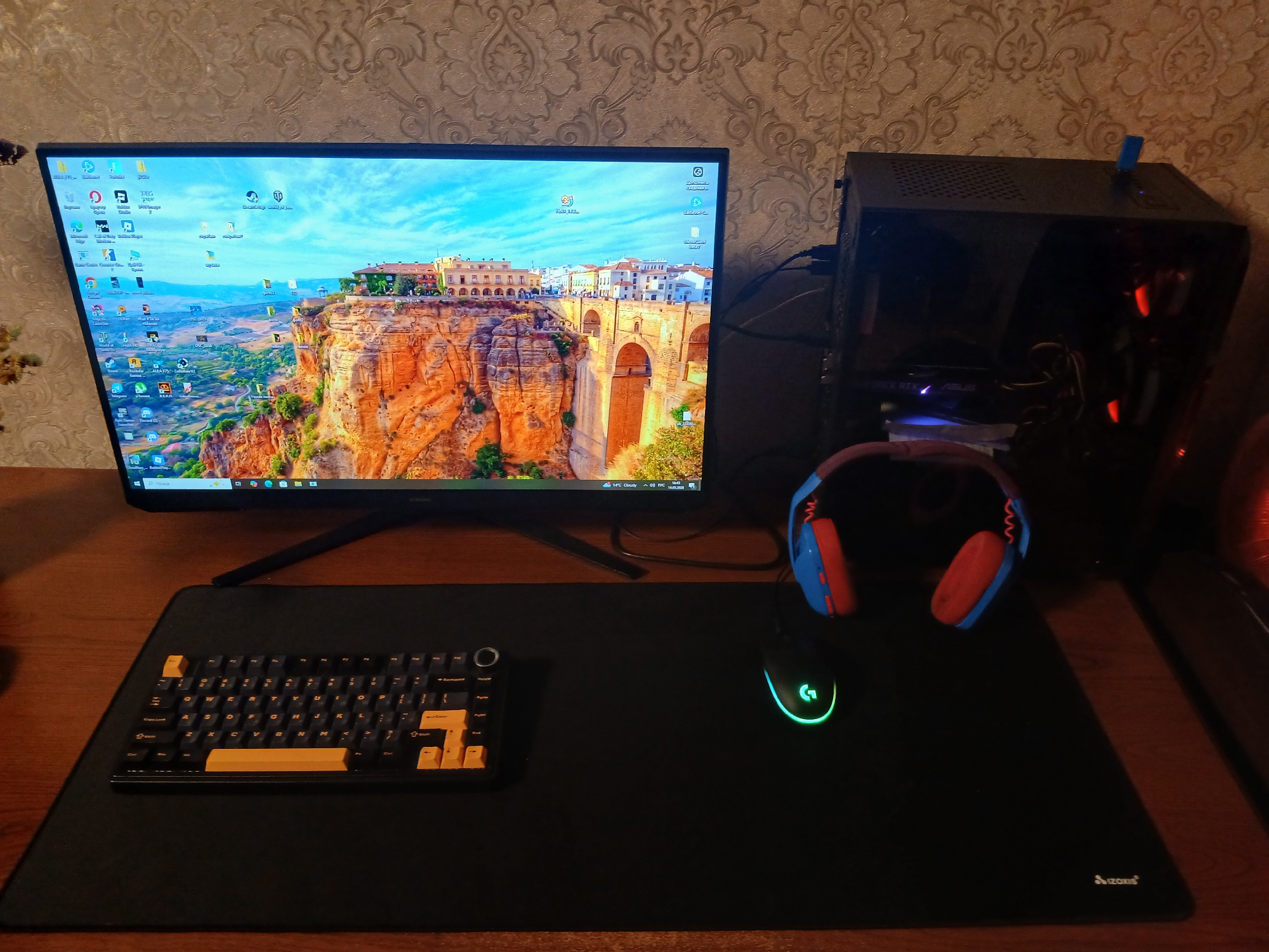 A desk setup featuring a monitor displaying a scenic view of Ronda, Spain, complete with a bridge and surrounding landscape. Adjacent to the monitor is a black desktop computer tower. On the desk is a black mousepad, with a custom mechanical keyboard featuring black and yellow keycaps, a Logitech gaming mouse with green LED lighting, and a blue and red gaming headset. The desk is wooden and the background features wallpaper with a subtle floral pattern. This image does not contain any specific indicators to suggest that it is located in the city of Kamianske, Ukraine.