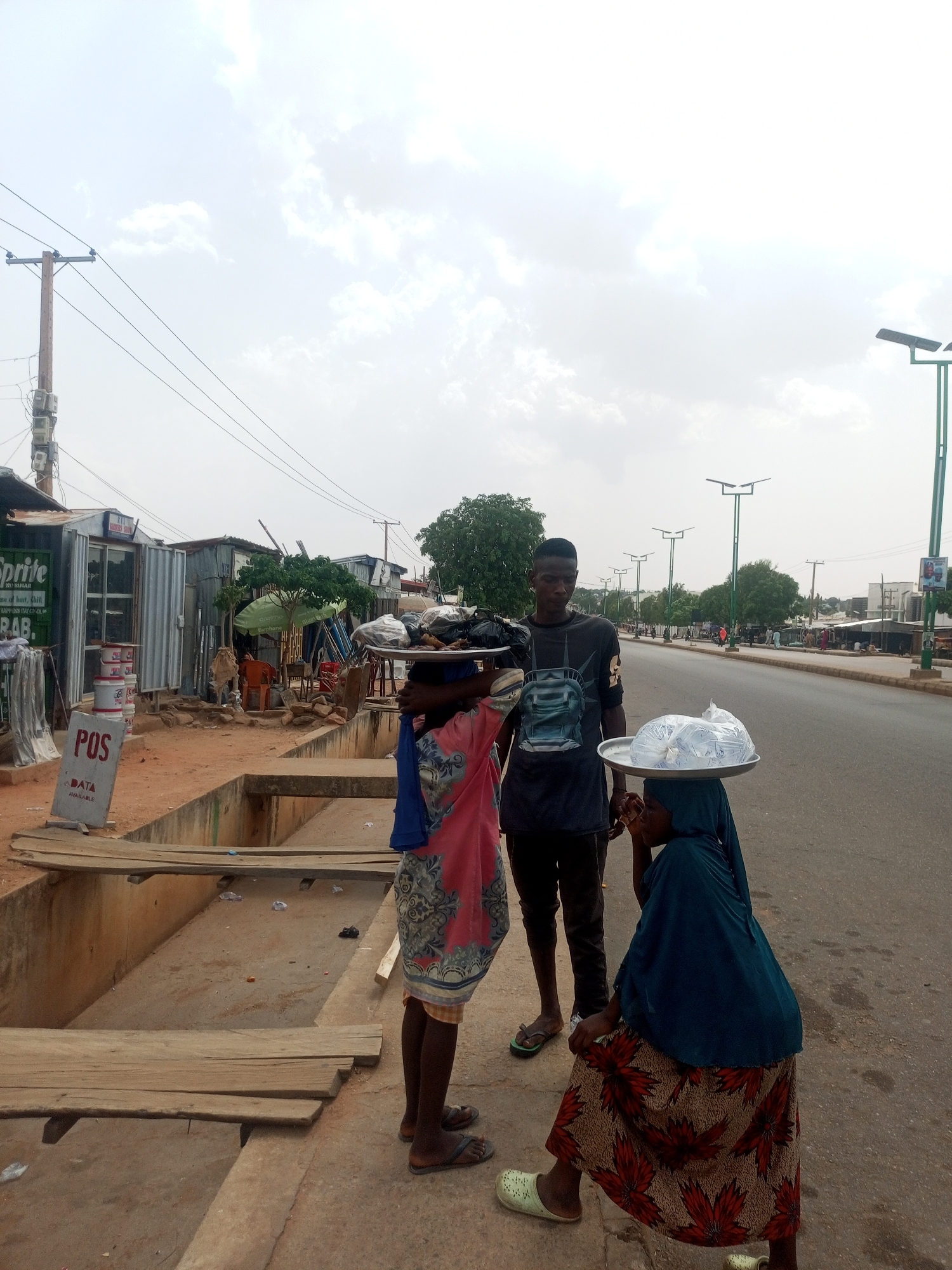 Here is a detailed description of the image:
**Scene Overview:** The image captures an outdoor scene in a city, potentially in a developing region. It features three people positioned beside a road or street. The setting appears to be a commercial area with buildings and infrastructure visible in the background.
**People:**
- Two individuals, seemingly young women, are carrying trays filled with items on their heads. One is wearing a pink patterned dress with a blue head covering, while the other has a dark green head covering and a patterned skirt.
- A young man stands between them, wearing a dark t-shirt and pants.
**Objects:**
- Trays: Each woman carries a tray on her head. One tray seems to contain bags of water, while the contents of the other are obscured.
- Infrastructure: The scene includes a road, a concrete sidewalk or curb, and an open drainage ditch covered with wooden planks for pedestrians to cross.
- Buildings: In the background, there are simple structures, possibly small shops or stalls. Some have corrugated metal walls. A green Sprite sign is visible on one of the buildings.
- Electrical poles: Electrical poles with wires can be seen extending across the scene.
- Streetlights: Modern streetlights are present along the road.
**Actions and Interactions:**
- The three people are standing together, possibly engaged in a transaction or conversation. The man appears to be interacting with one of the women.
- The women carrying trays suggest they are vendors or sellers, transporting goods for sale.
**Environment:**
- The sky is overcast with a light, cloudy atmosphere.
- The surroundings suggest a modest or lower-income area, with simple buildings and infrastructure.
- The road appears relatively quiet, with few vehicles visible.
**Location Details:** The exact location is given as the city of Unknown, Unknown.