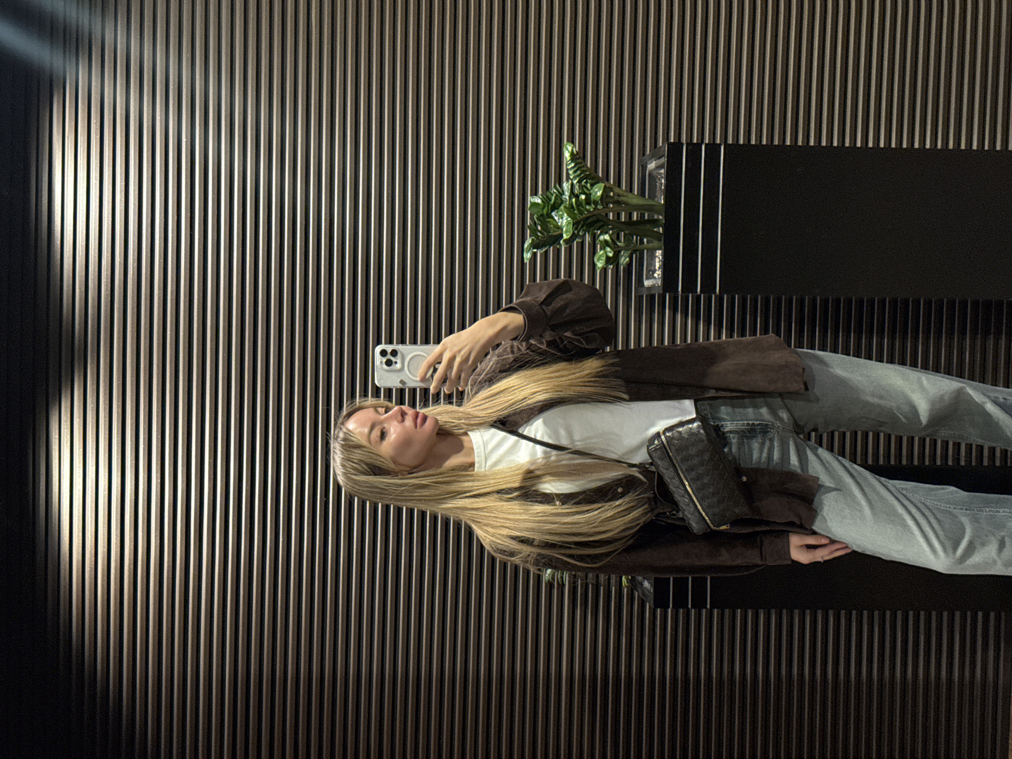 A woman stands in front of a wall made of vertical wooden slats in what appears to be an indoor setting, possibly a public space. The woman is dressed in a white t-shirt, light blue jeans, and a brown jacket. She is holding a white smartphone and appears to be taking a picture or video. A black crossbody bag with a woven pattern hangs across her body. To the right of the woman is a black rectangular planter with green plants. The location may be in Moscow, Russia.