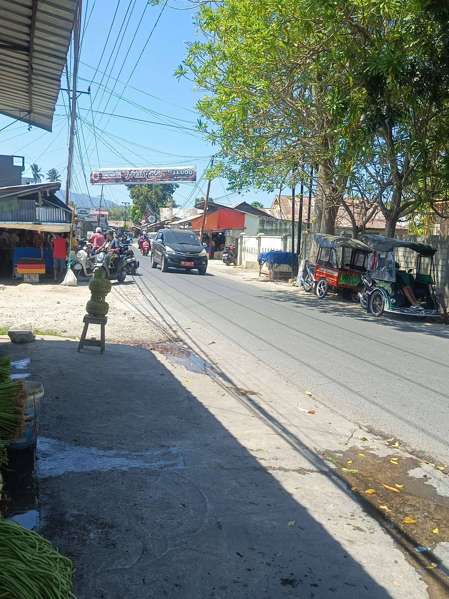 A bright, sunny daytime scene depicts a street in Tenggela, Indonesia, with a road running through it. On the left side of the road, a covered market stall is visible, with several people standing and interacting. A black car with license plate "DM 1153 P" is driving on the road. Further down the street, a trike-like vehicle is parked. In the foreground, bundles of green vegetables are stacked in buckets. The sky is clear and blue, with a large tree on the right casting a shadow across the scene. The general atmosphere is that of a busy marketplace in a tropical climate.