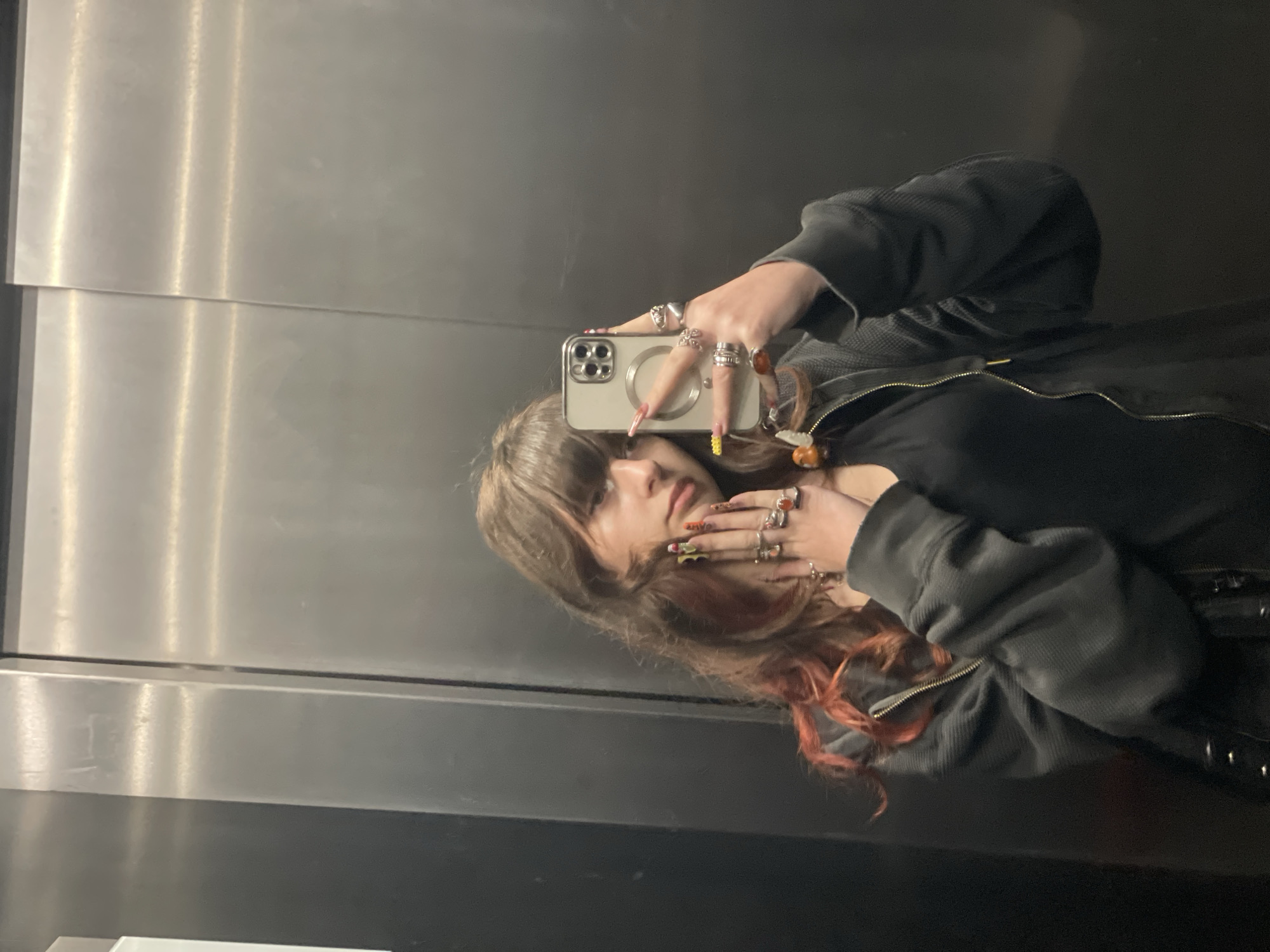 A young woman takes a selfie in what appears to be an elevator with brushed metal walls. She is wearing a dark top under a gray textured jacket with a zipper. She has medium-length brown hair with reddish-orange highlights. Multiple rings adorn her fingers, and she holds a light-colored smartphone in front of her face to take the photo. Due to the lack of distinctive architectural or environmental features, it is impossible to determine if the location is in Raanana, Israel.
