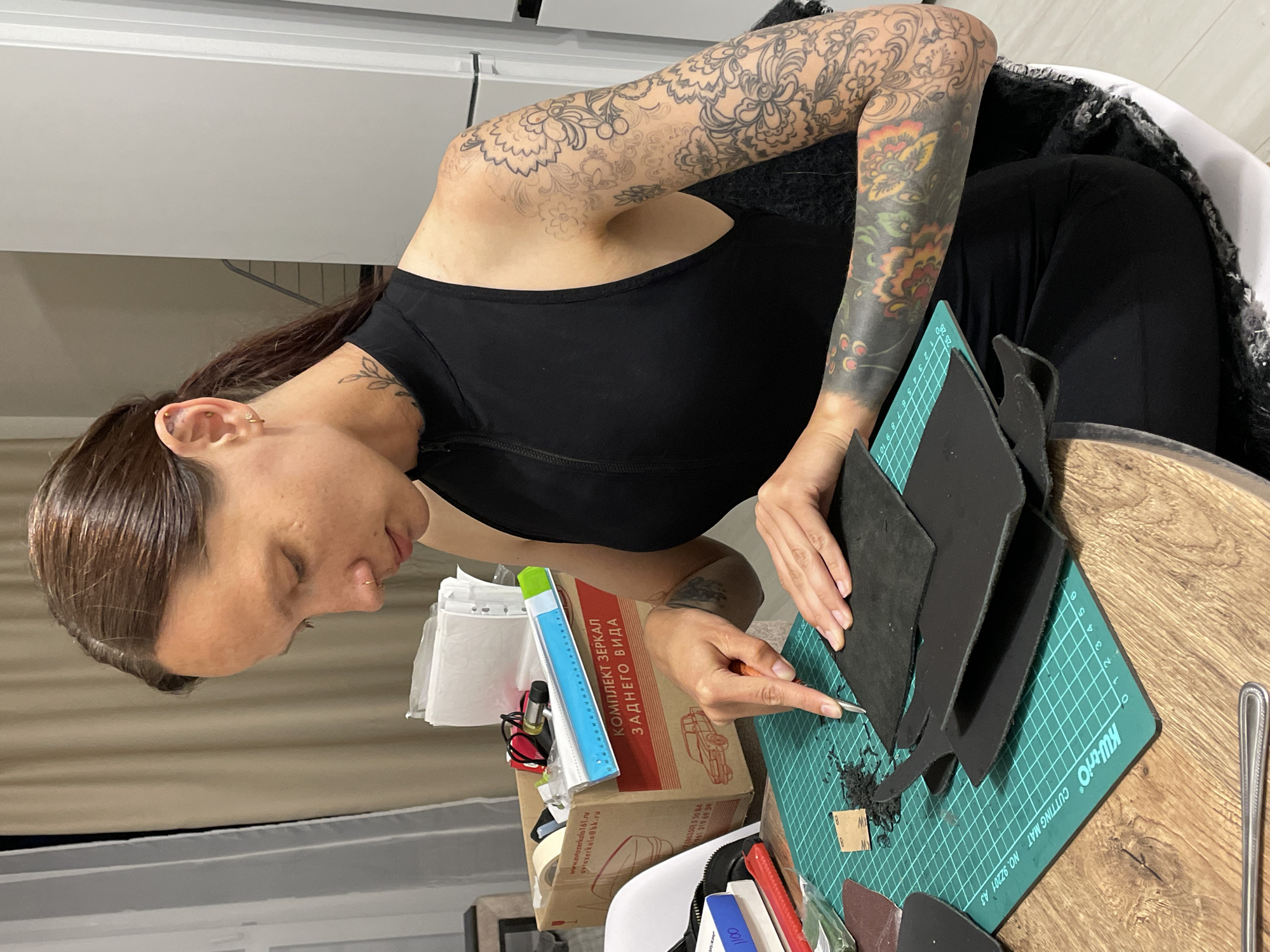A woman with dark hair pulled back in a ponytail and multiple tattoos on her arms is working on a craft project at a wooden table. She is wearing a black sleeveless top. In front of her is a green cutting mat with grid lines, on which several pieces of black material are placed. She is using a craft knife to cut one of the pieces. To her left is a cardboard box with text in Russian, containing various crafting supplies, including a blue ruler. The background shows a curtained window and a white appliance, possibly a refrigerator. The scene appears to be an indoor setting, likely a home or studio, in the city of Pervomaysky, Russia.