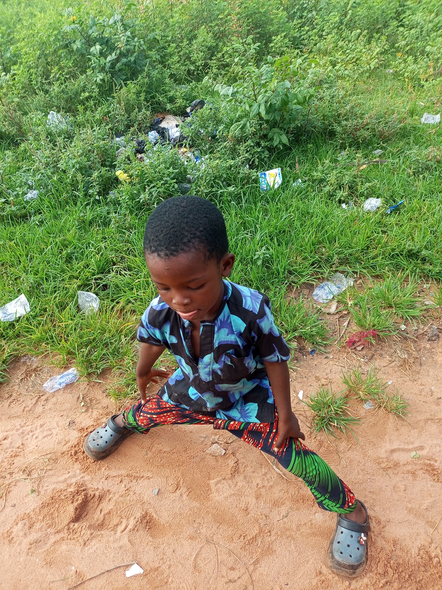 A young Black male child sits on sandy ground in front of a dense green embankment covered in weeds and litter, including plastic bottles and wrappers. The child is wearing a short-sleeved shirt with a blue, black, and green floral pattern, and pants with a red, black, and green geometric pattern. He has black hair and is looking down with his tongue slightly out. He is wearing dark grey Crocs on both feet. The scene appears to be an outdoor area, possibly on the outskirts of Tempure, Nigeria, given the arid ground and sparse vegetation.